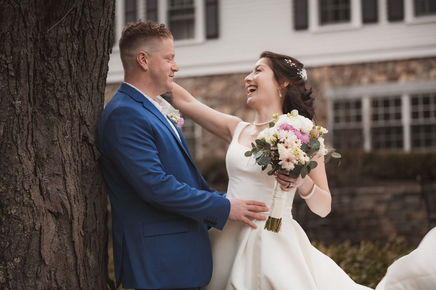 Laughing bride in white gown with bouquet touches groom's face as he leans against tree at outdoor wedding venue.