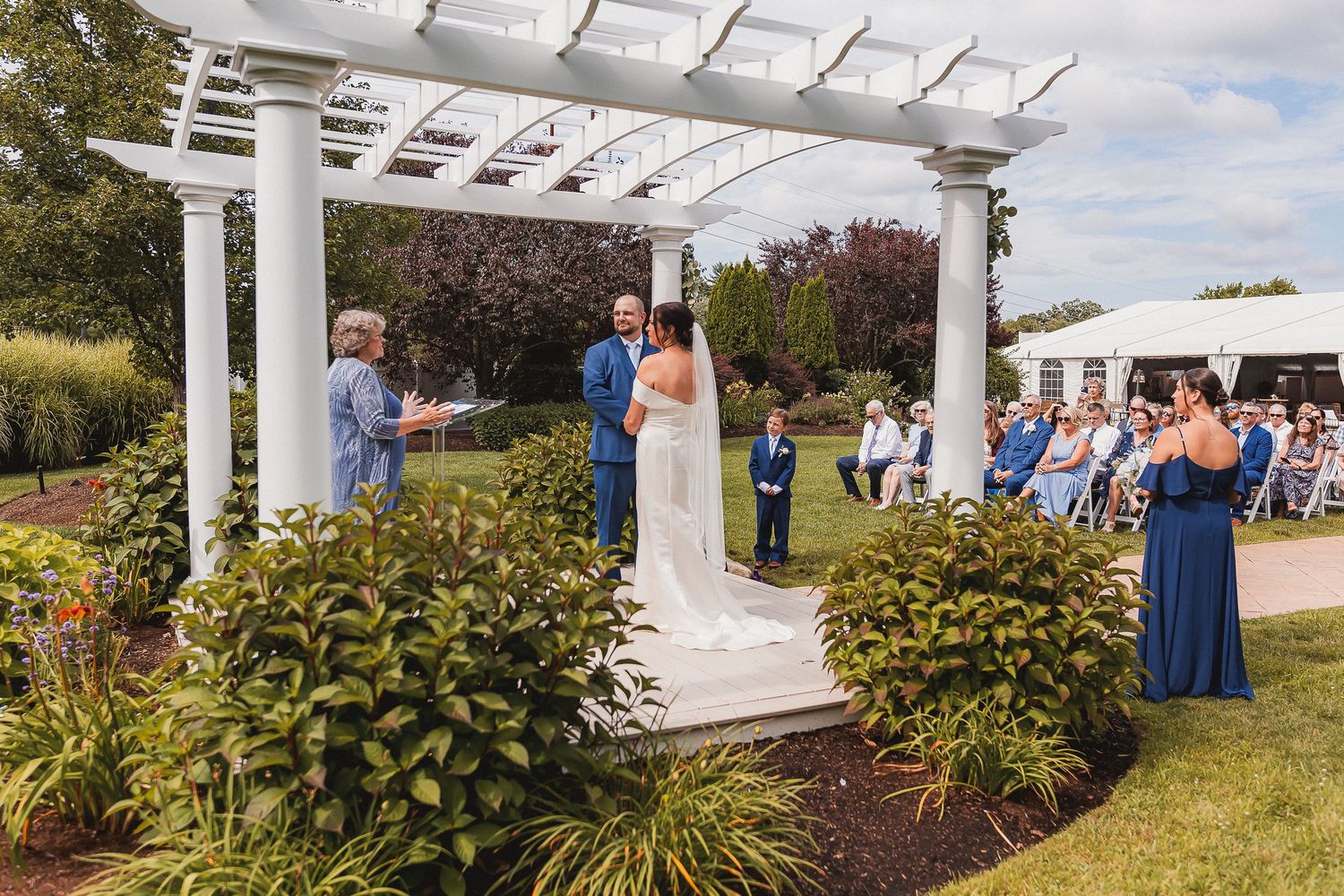 Bride and groom exchanging vows under white pergola with seated guests and blue bridesmaids outdoors.