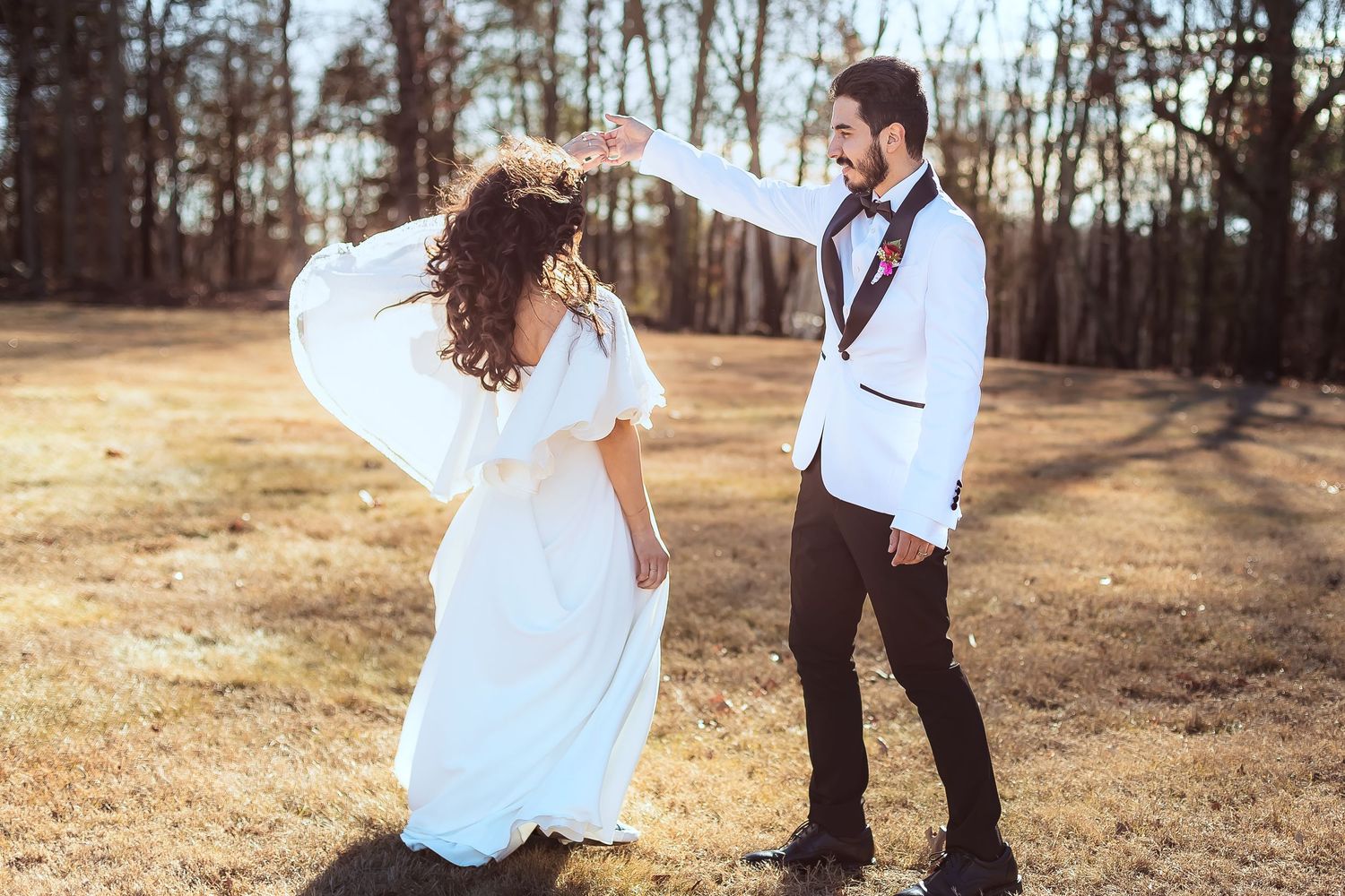 Bride and groom dancing playfully in sunlit field, groom in white jacket twirling bride.