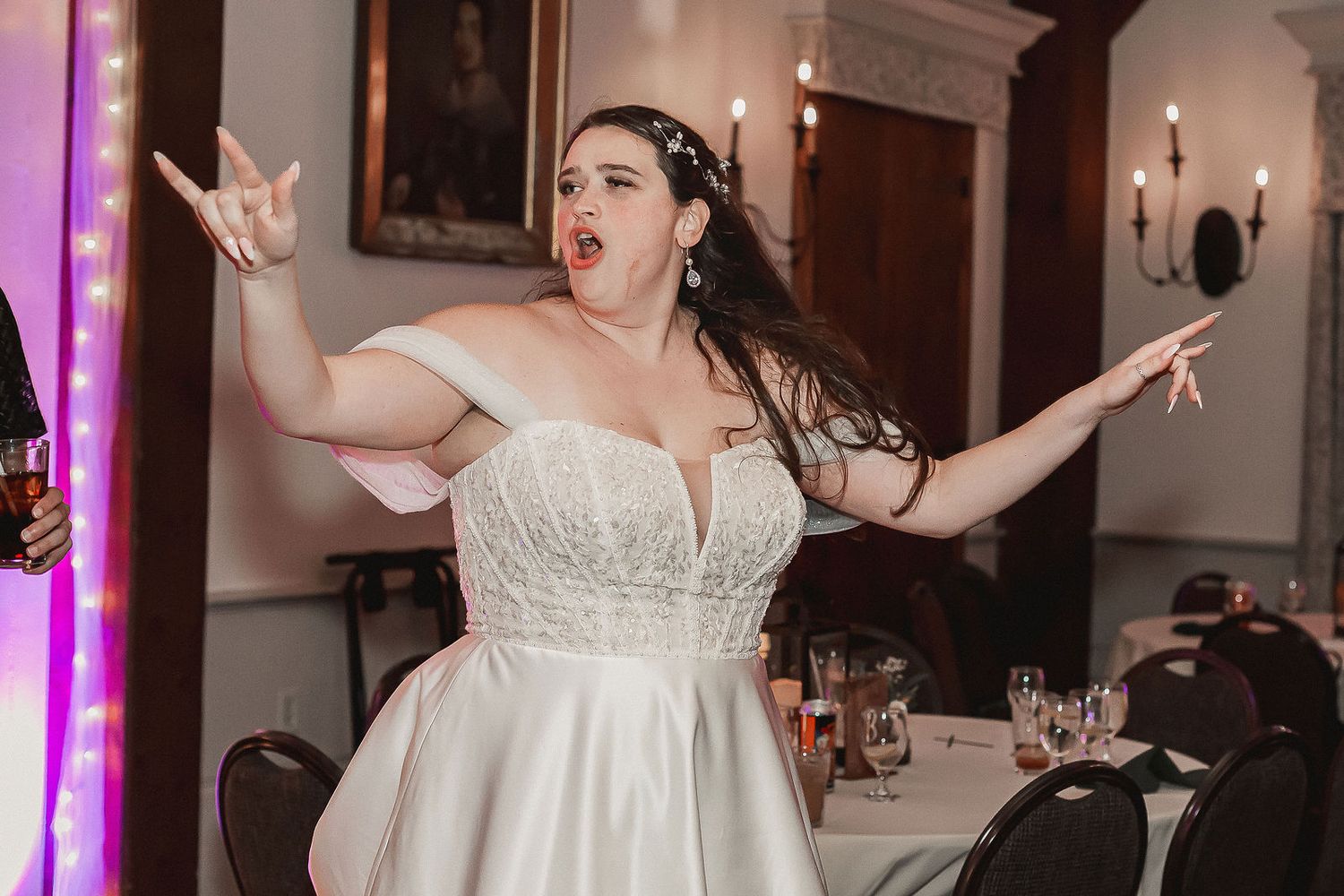 Excited bride in off-shoulder white wedding gown celebrates with arms outstretched at reception.