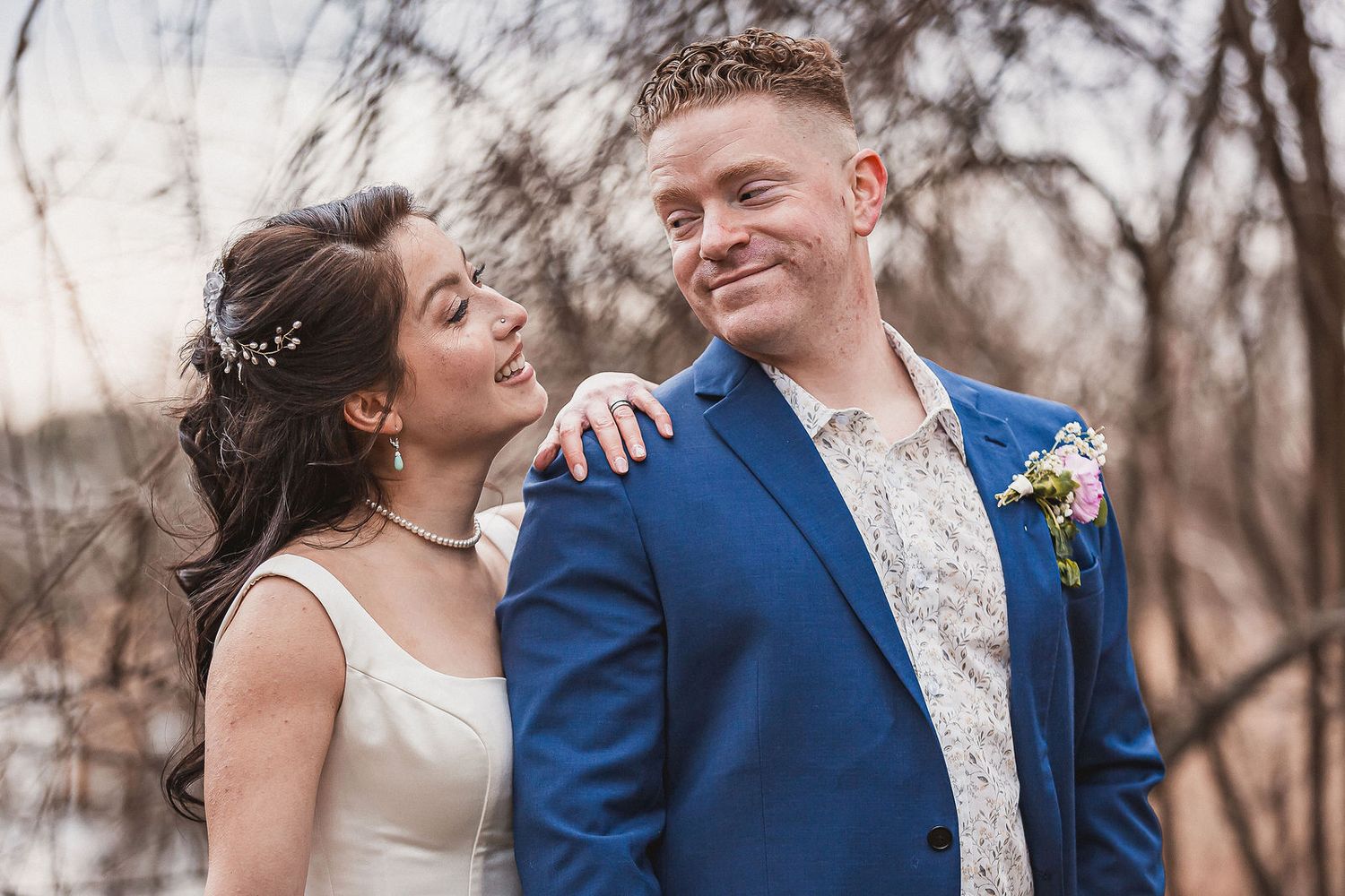 Bride and groom smiling outdoors, groom in blue suit with floral boutonniere at sunset.