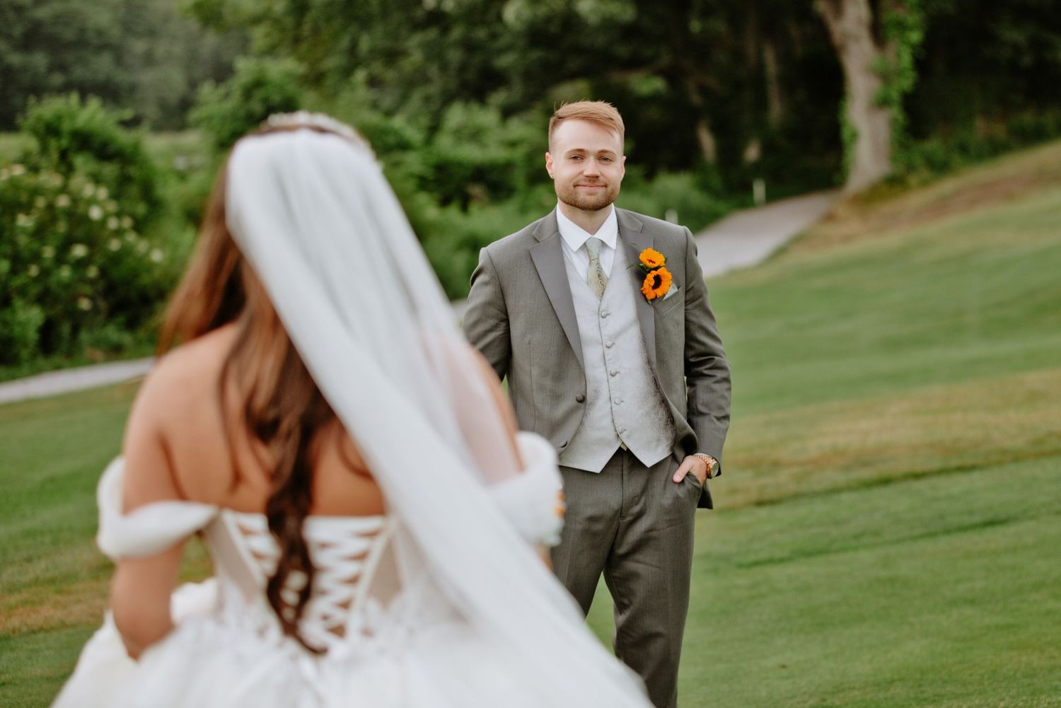 Groom in gray suit smiling at bride in white corset gown during outdoor first look on golf course.