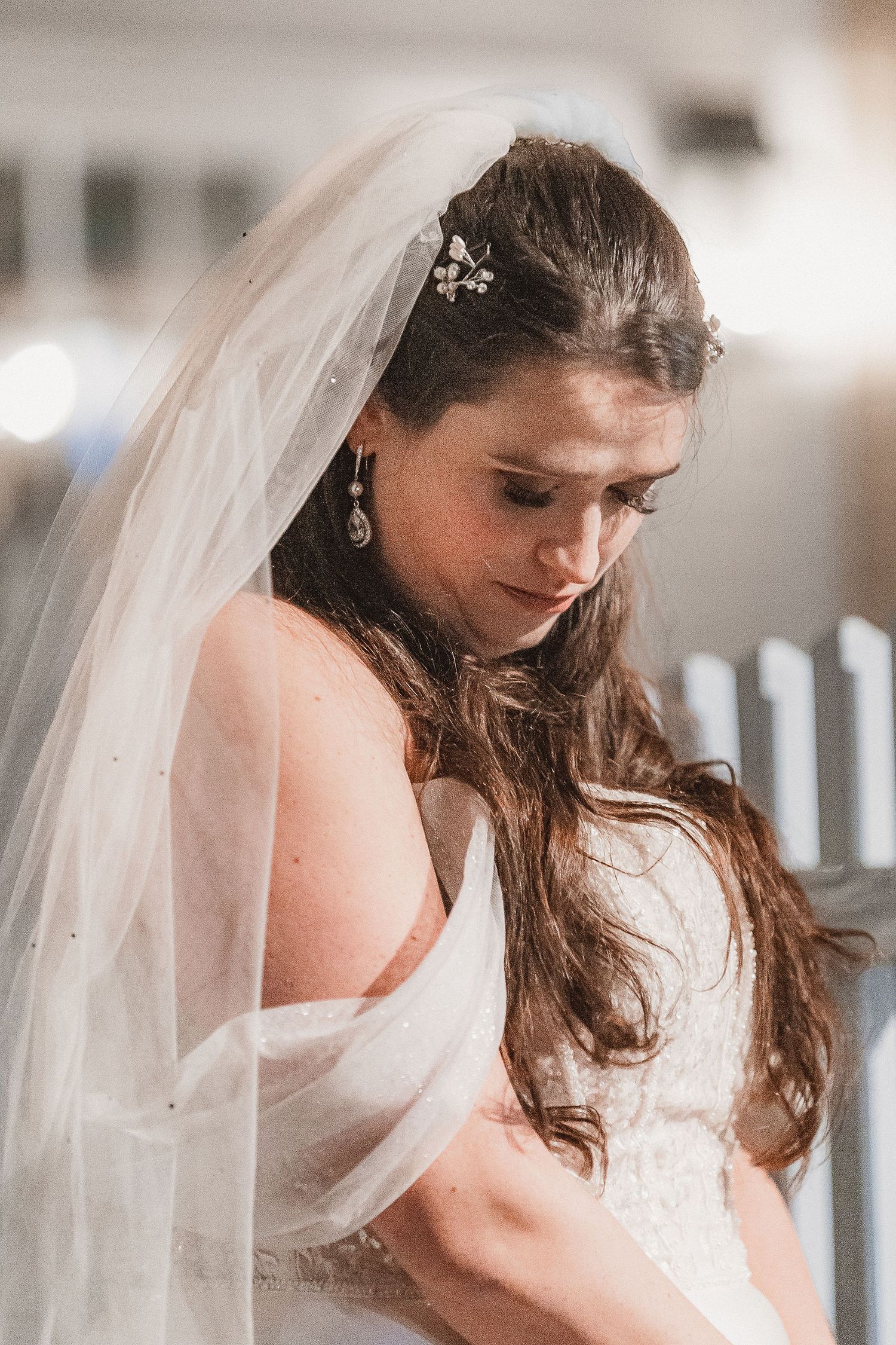 Beautiful bride in a lace gown and veil with floral hair accessories looking down.