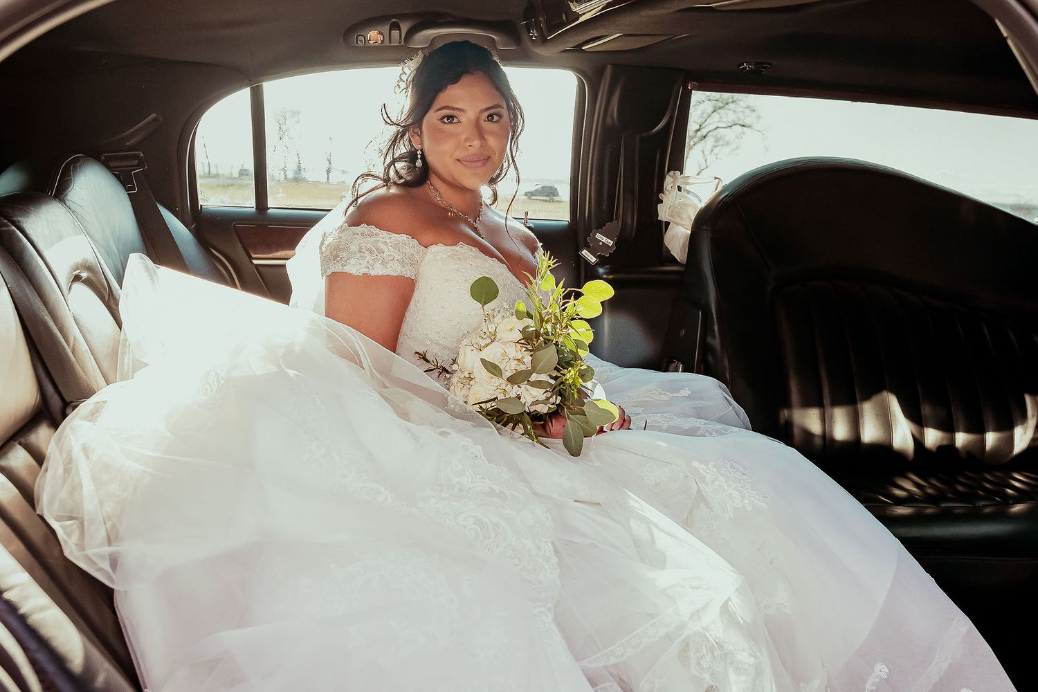 Bride in white ball gown holding bouquet seated in limousine on wedding day.