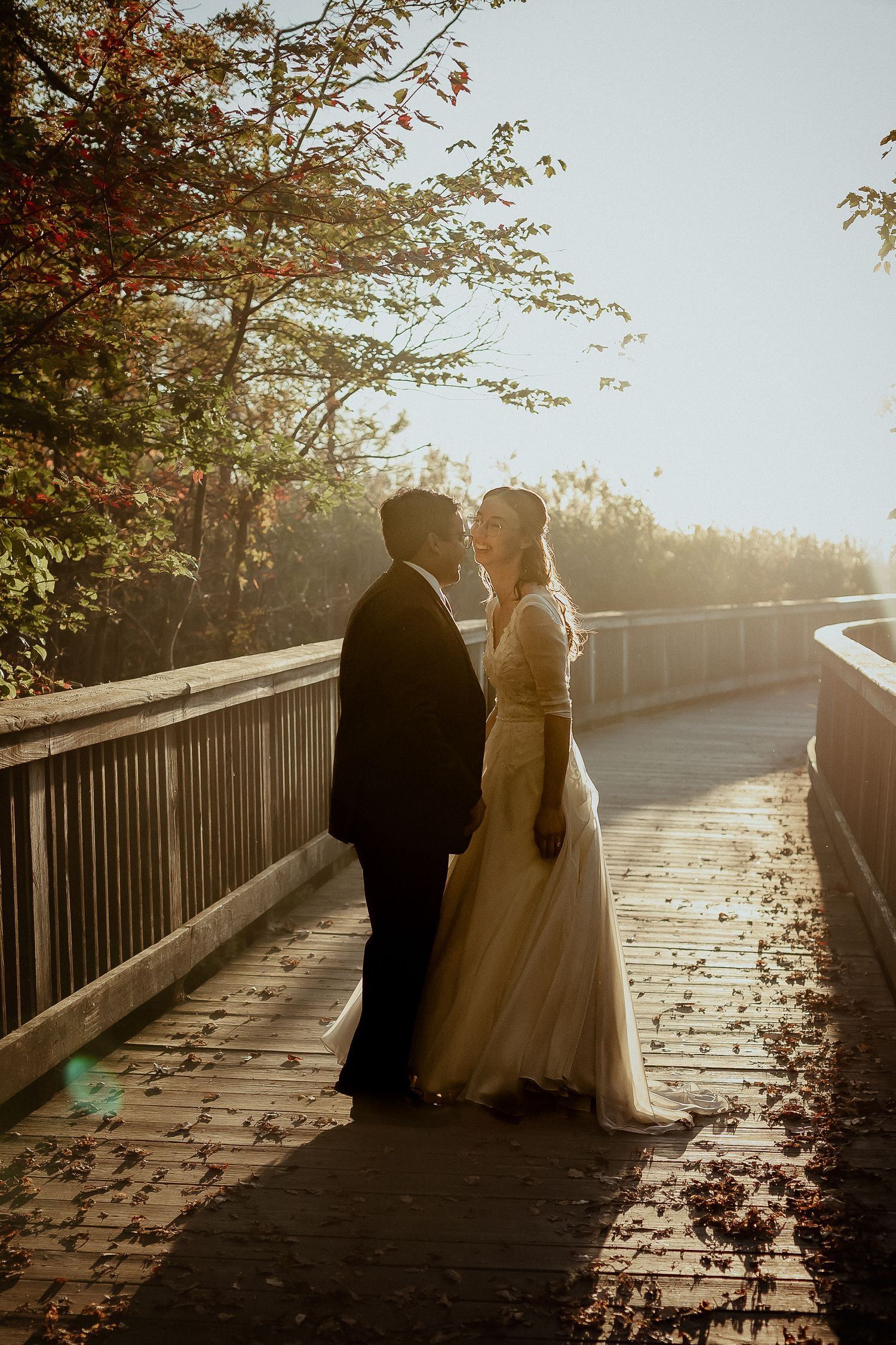 Bride and groom share a kiss on a sunlit wooden bridge surrounded by autumn foliage.