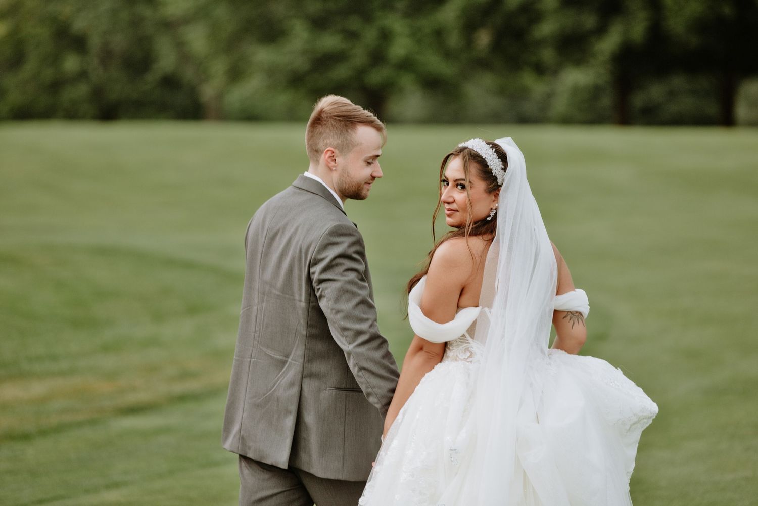Bride in white off-shoulder gown with veil looks back at groom in gray suit on green lawn.