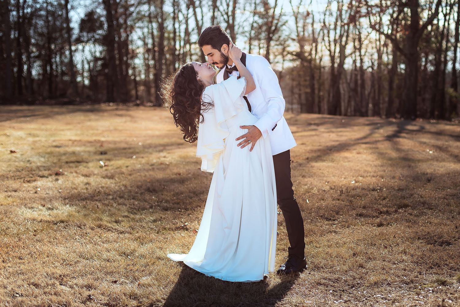 Bride and groom sharing romantic kiss outdoors in sunlit field with bare winter trees behind them.