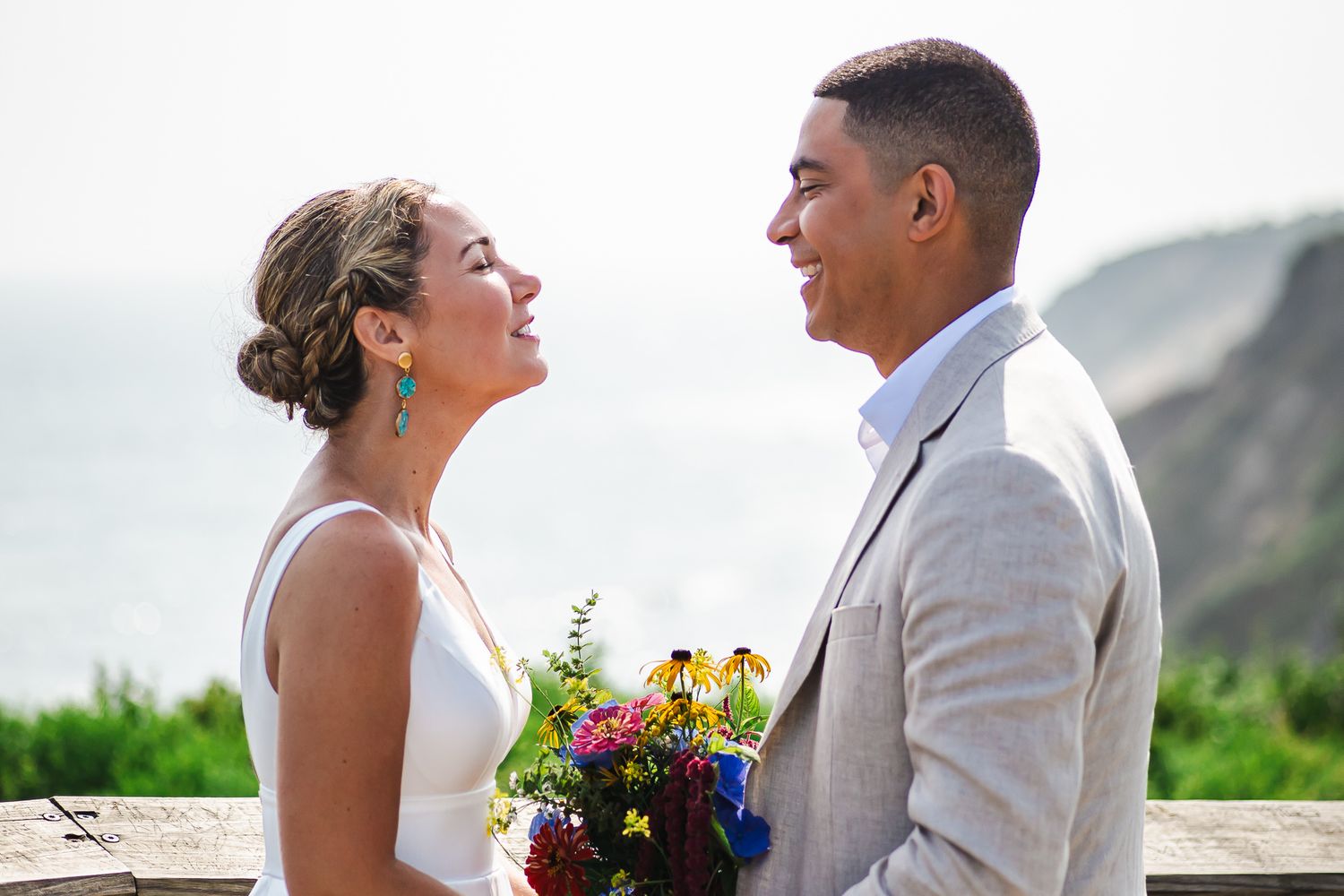 Smiling bride and groom in beige suit share a joyful moment with ocean views in the background.