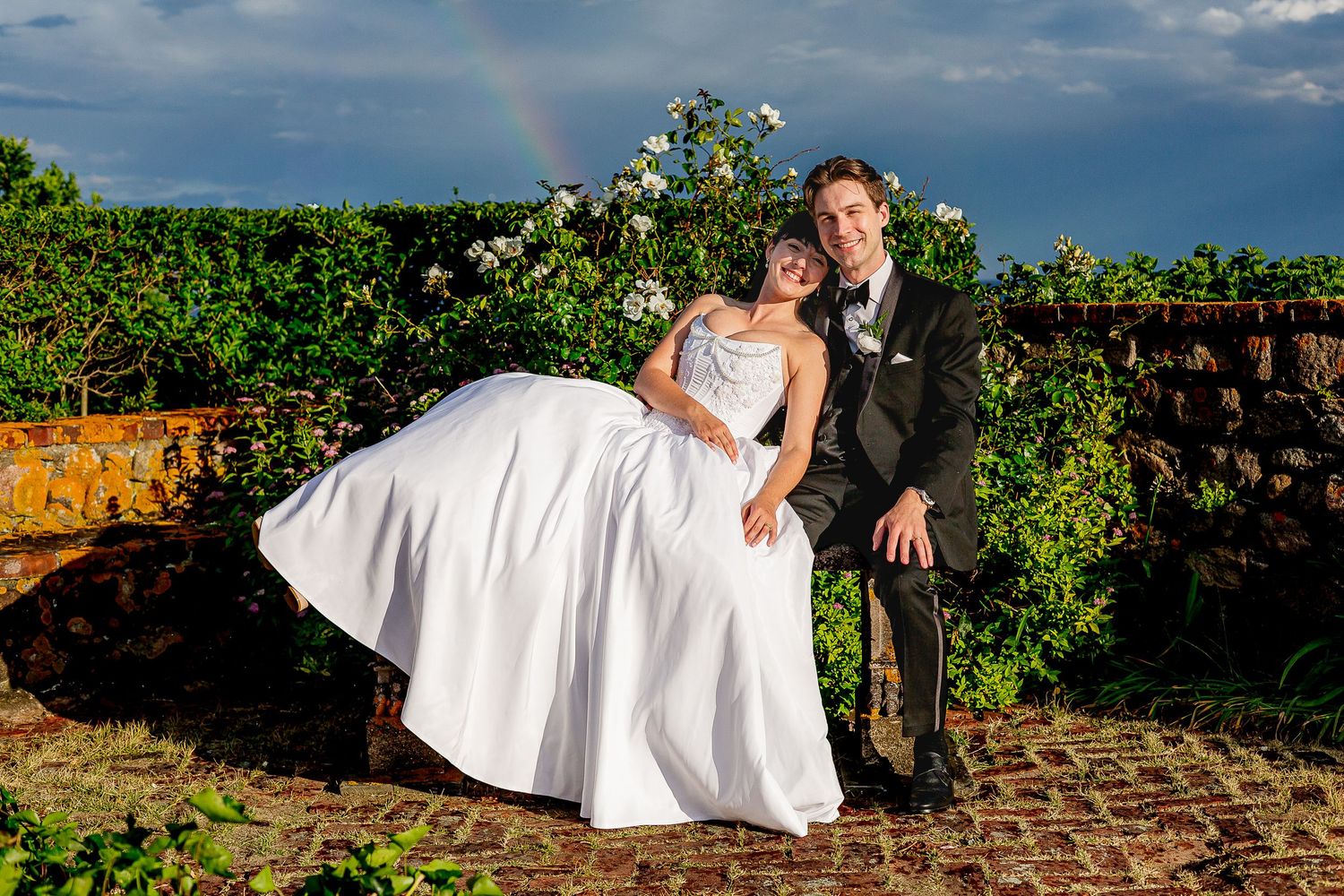 Bride and groom pose joyfully outdoors with a rainbow and blooming roses behind them on their wedding day.