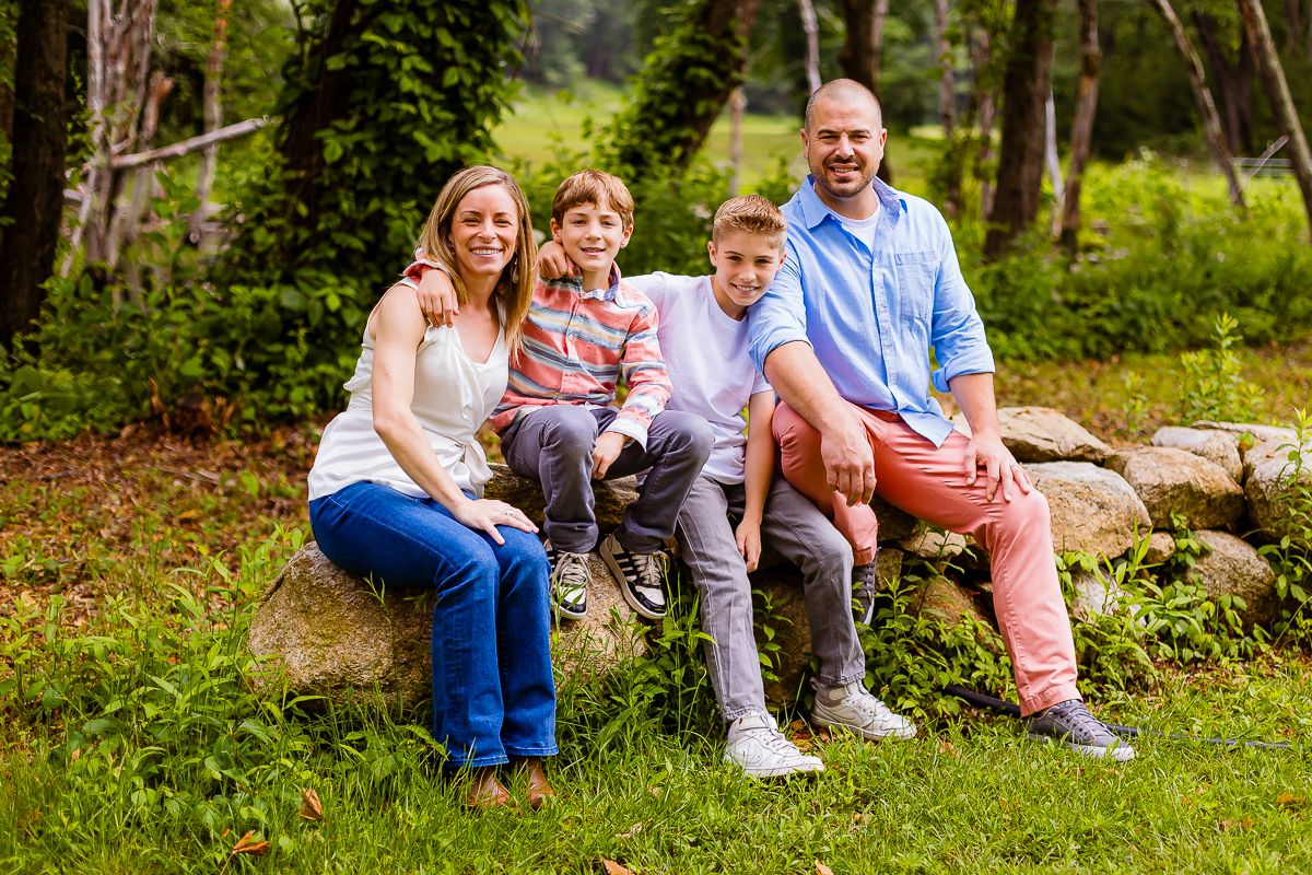Happy family of four sitting on large rock surrounded by lush green trees in outdoor nature portrait session.