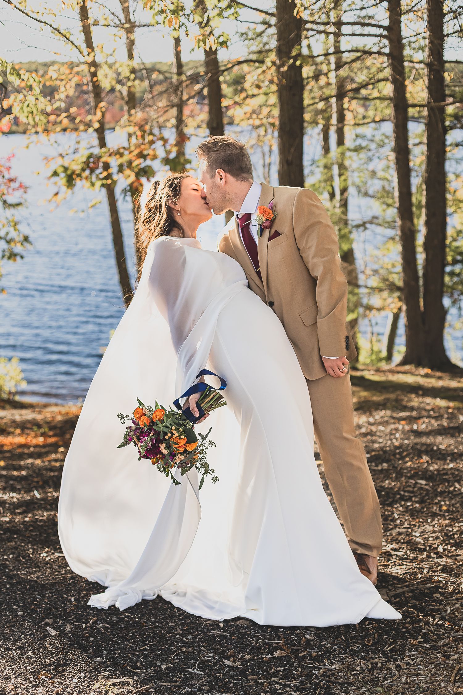 Bride and groom share a kiss near a sunlit lake in fall, bride holding a vibrant bouquet.