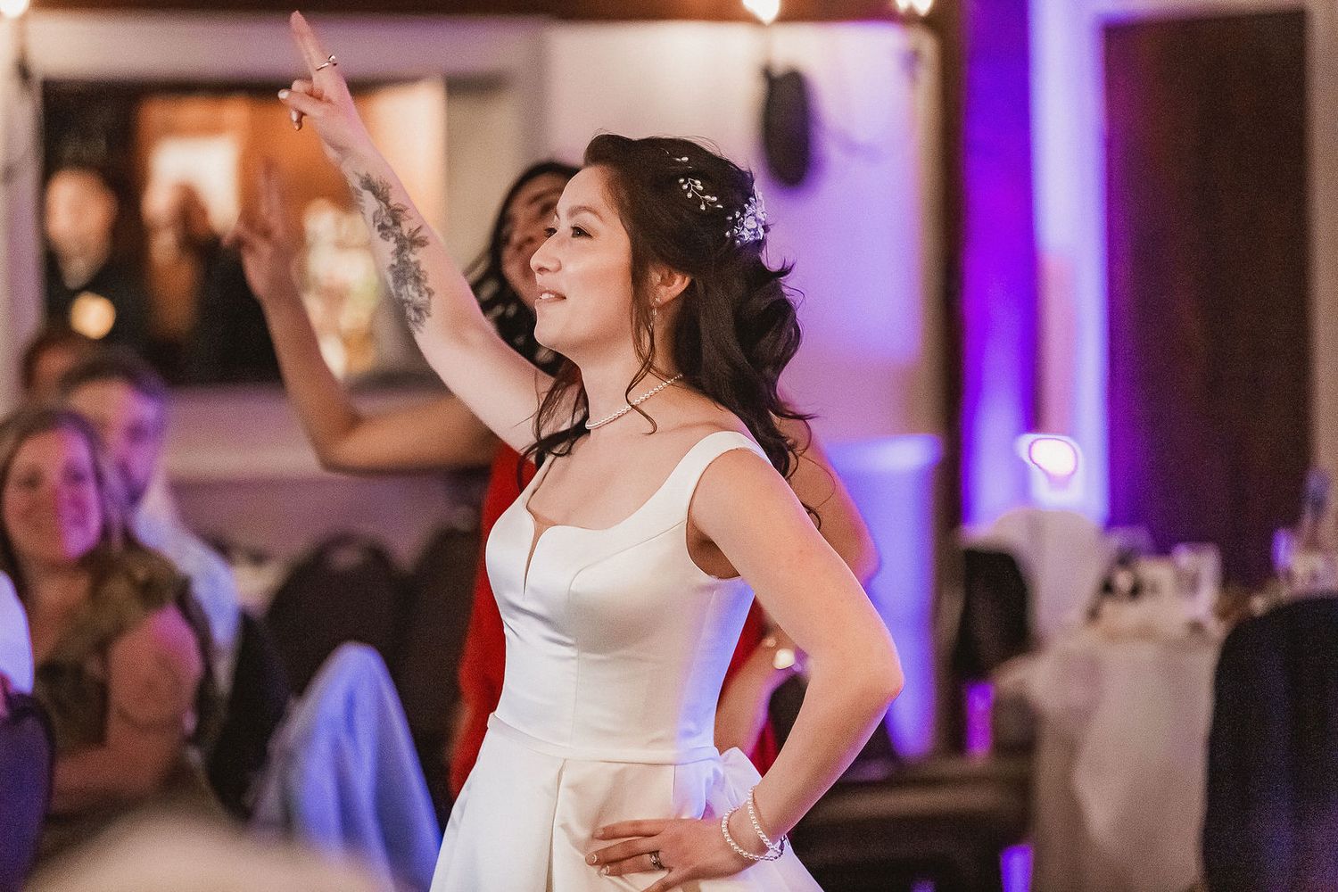 Bride in white gown pointing upward confidently during wedding reception celebration.