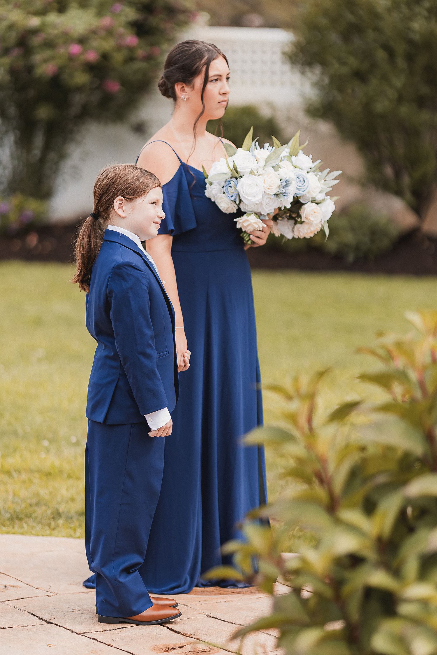 Bridesmaid in navy blue gown holding white floral bouquet standing with young boy in blue suit.