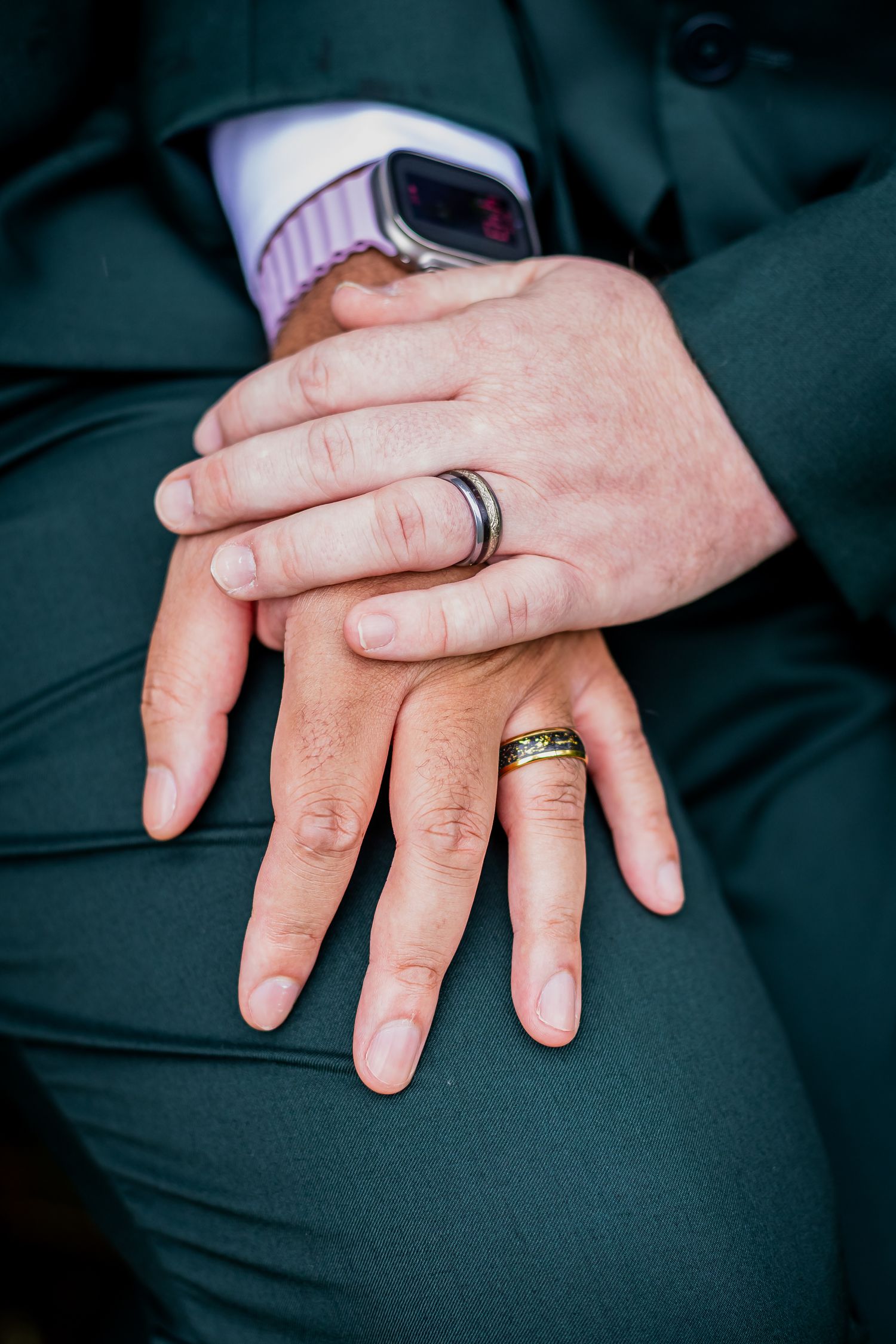 Close-up of two grooms hands folded together wearing wedding bands in green suits.