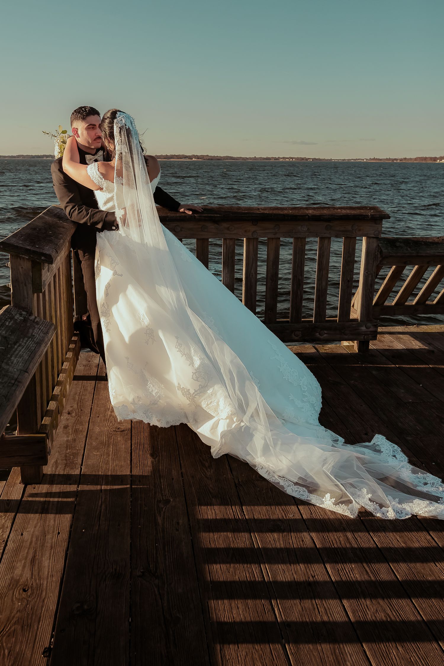 Bride and groom kissing on wooden dock with long lace veil trailing at sunset.