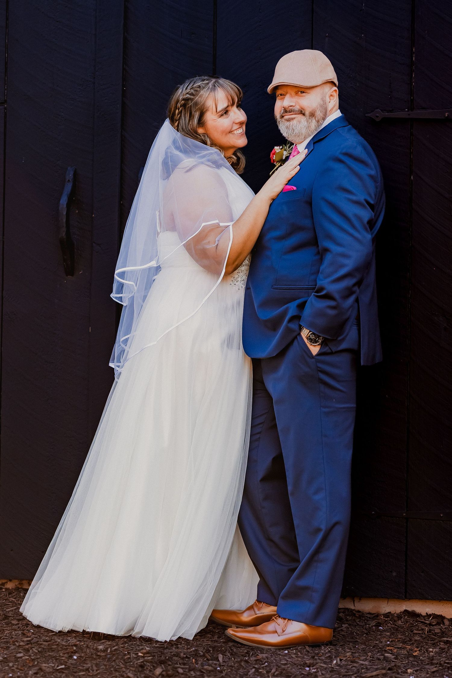 Bride in white gown and groom in blue suit posing together against a dark barn door.