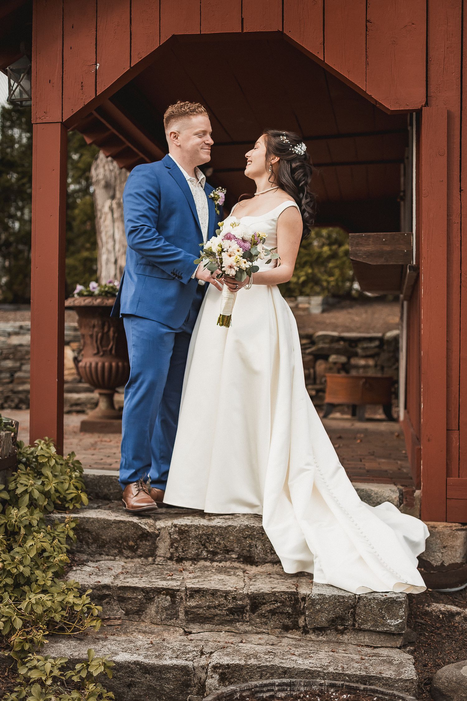 Bride and groom posing on stone steps beneath red wooden arch, bride holding colorful bouquet at rustic wedding venue.