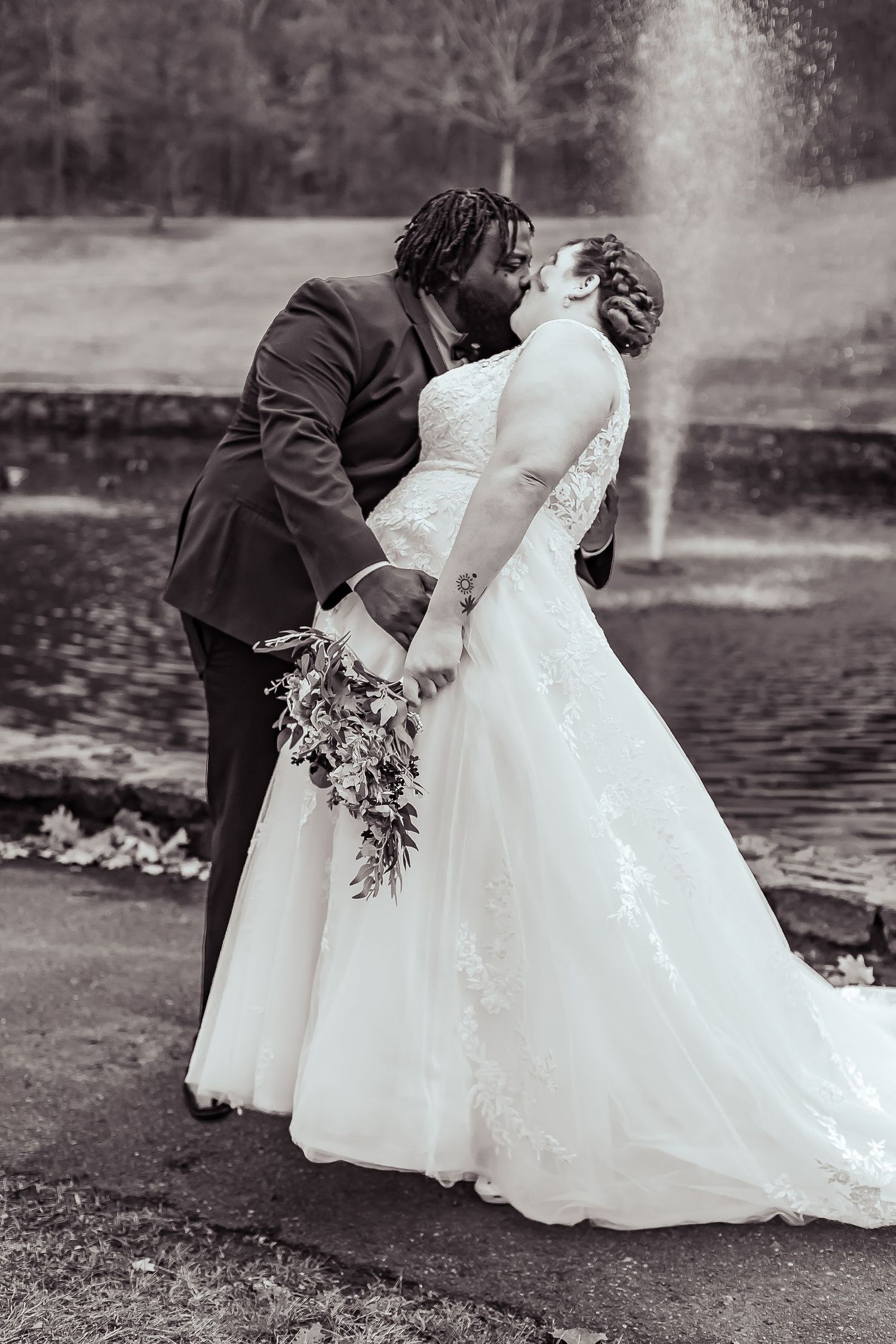 Black and white photo of interracial couple sharing wedding kiss near fountain with bridal bouquet.