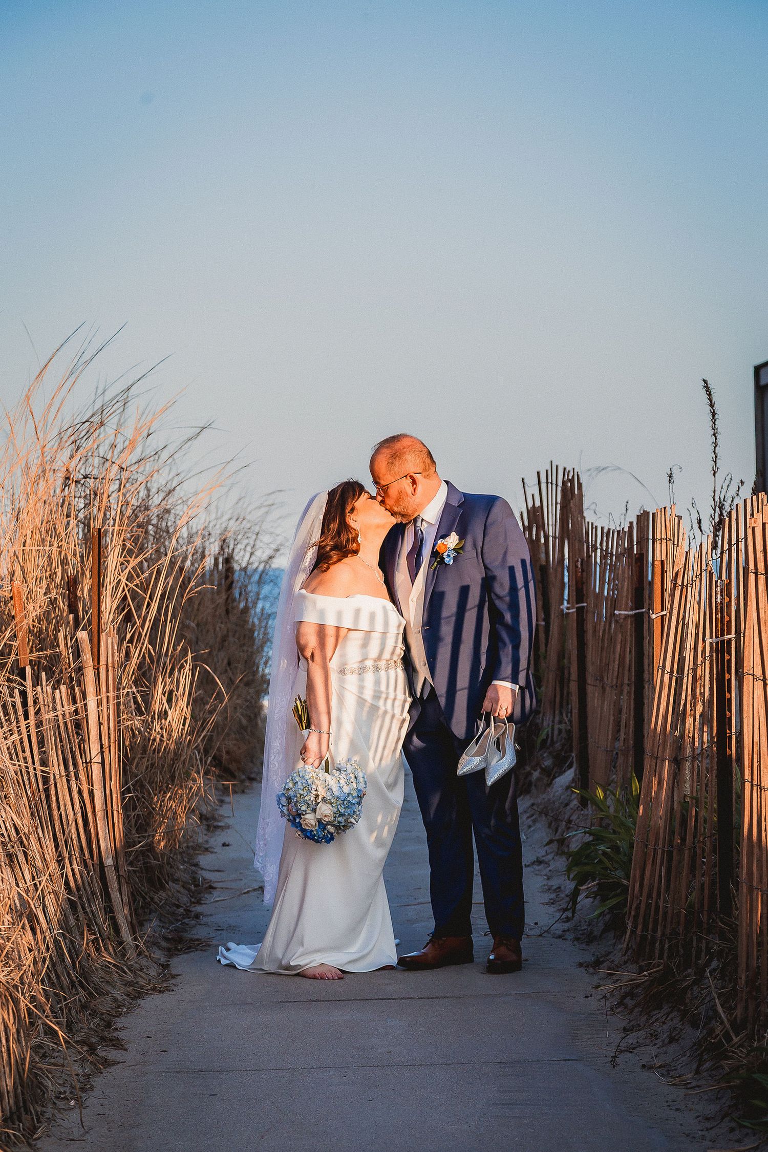 Bride and groom sharing romantic kiss on beach pathway at golden hour, bride holding blue bouquet.