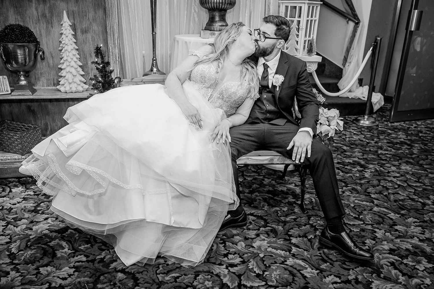 Black and white photo of bride leaning in to kiss groom seated in chair at elegant wedding venue.