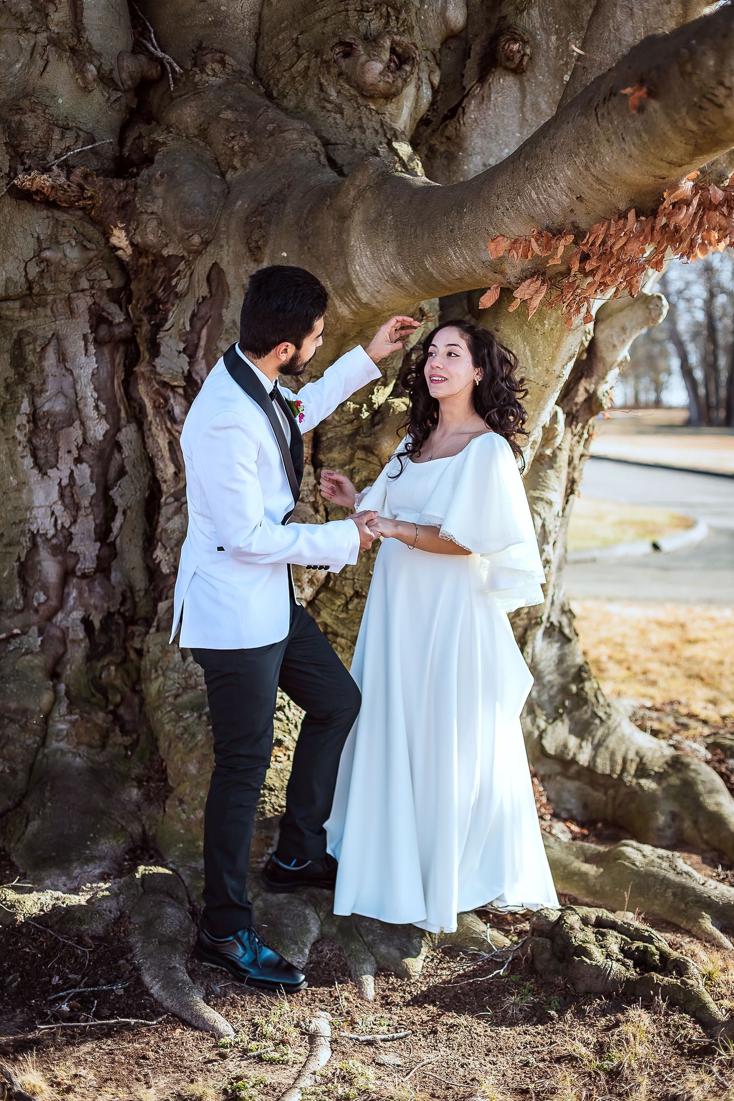 Bride and groom share a romantic moment under a large tree in winter wedding attire.