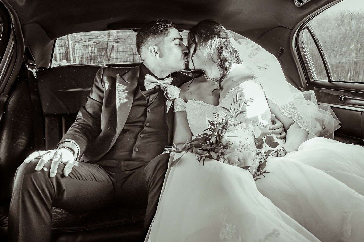 Newlyweds share a romantic kiss in the backseat of a car in black and white.
