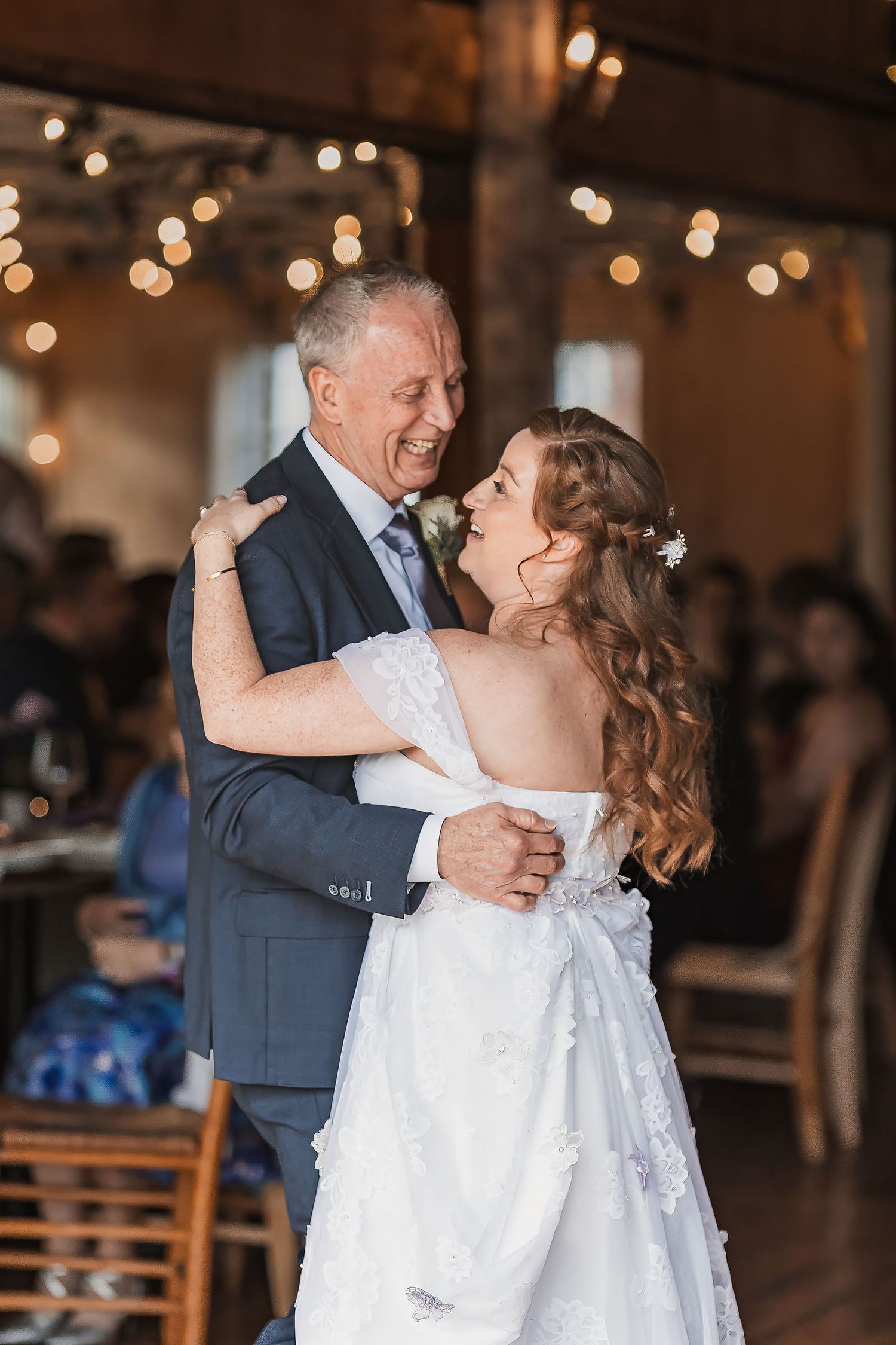 Bride sharing joyful father-daughter dance under romantic string lights at indoor wedding reception.