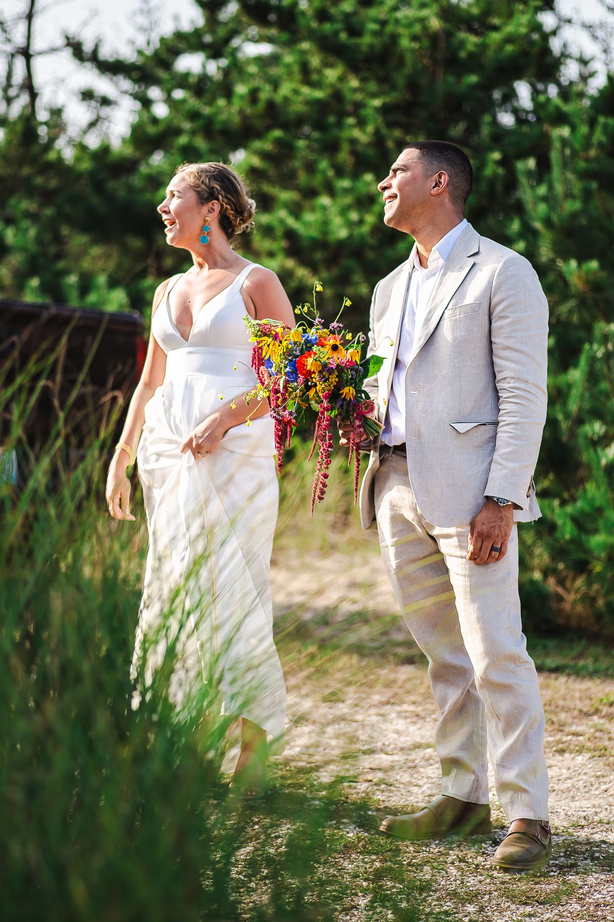 Joyful bride and groom walking outdoors, bride in white gown holding colorful bouquet, groom in beige suit.