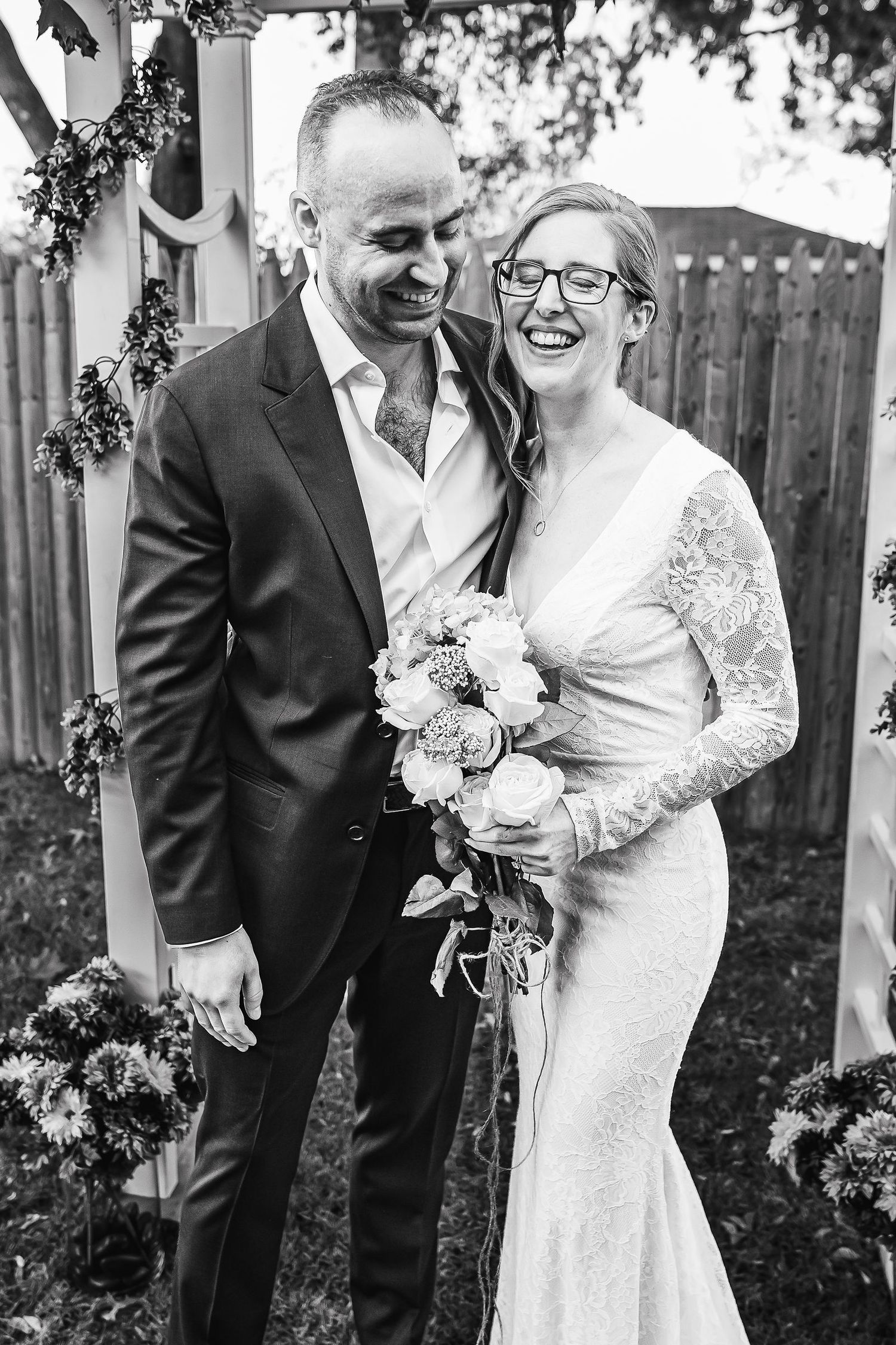 Happy bride wearing glasses holds bouquet while laughing with groom at outdoor wedding ceremony.