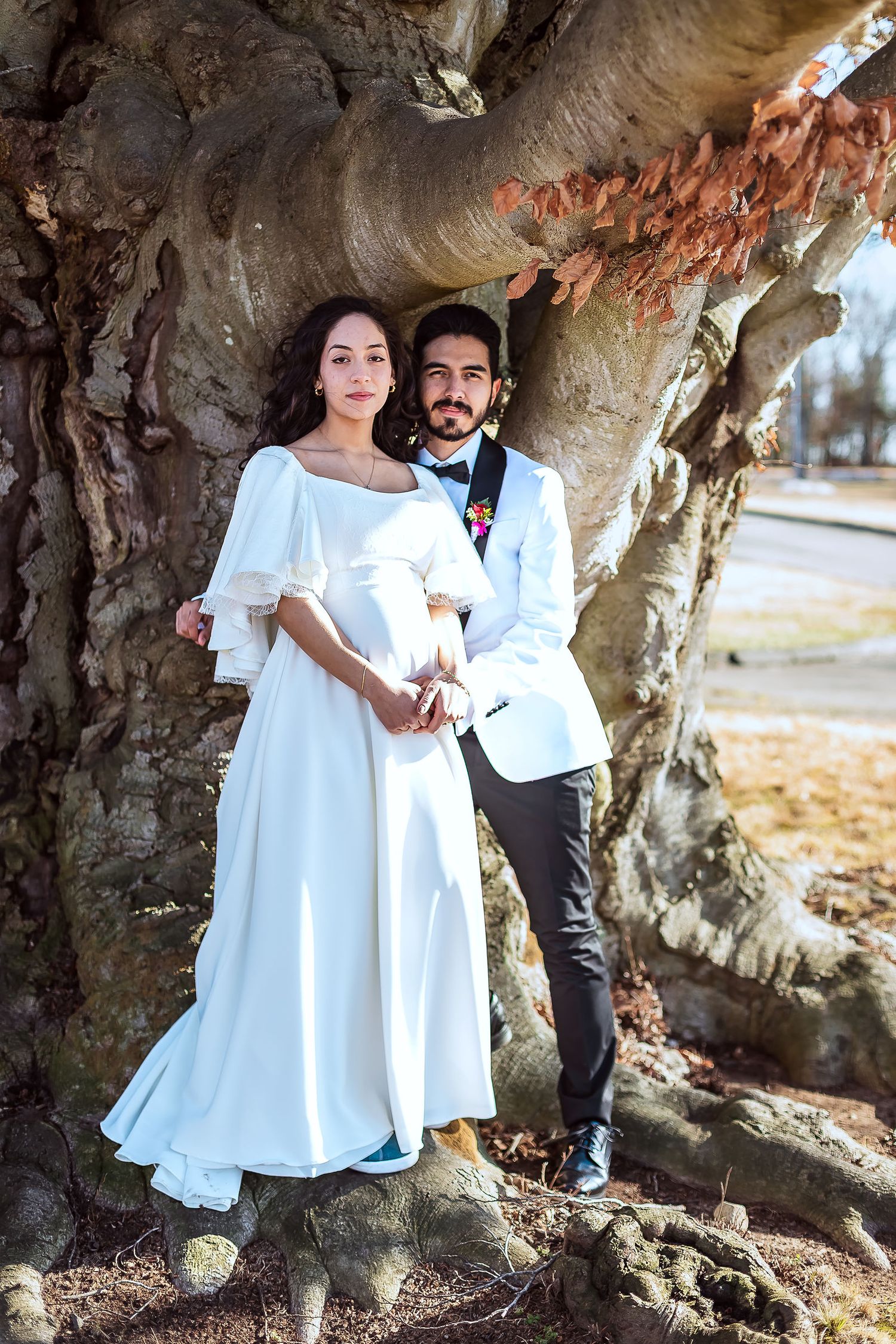 Bride in white gown and groom in white jacket posing together beside large tree with autumn leaves.
