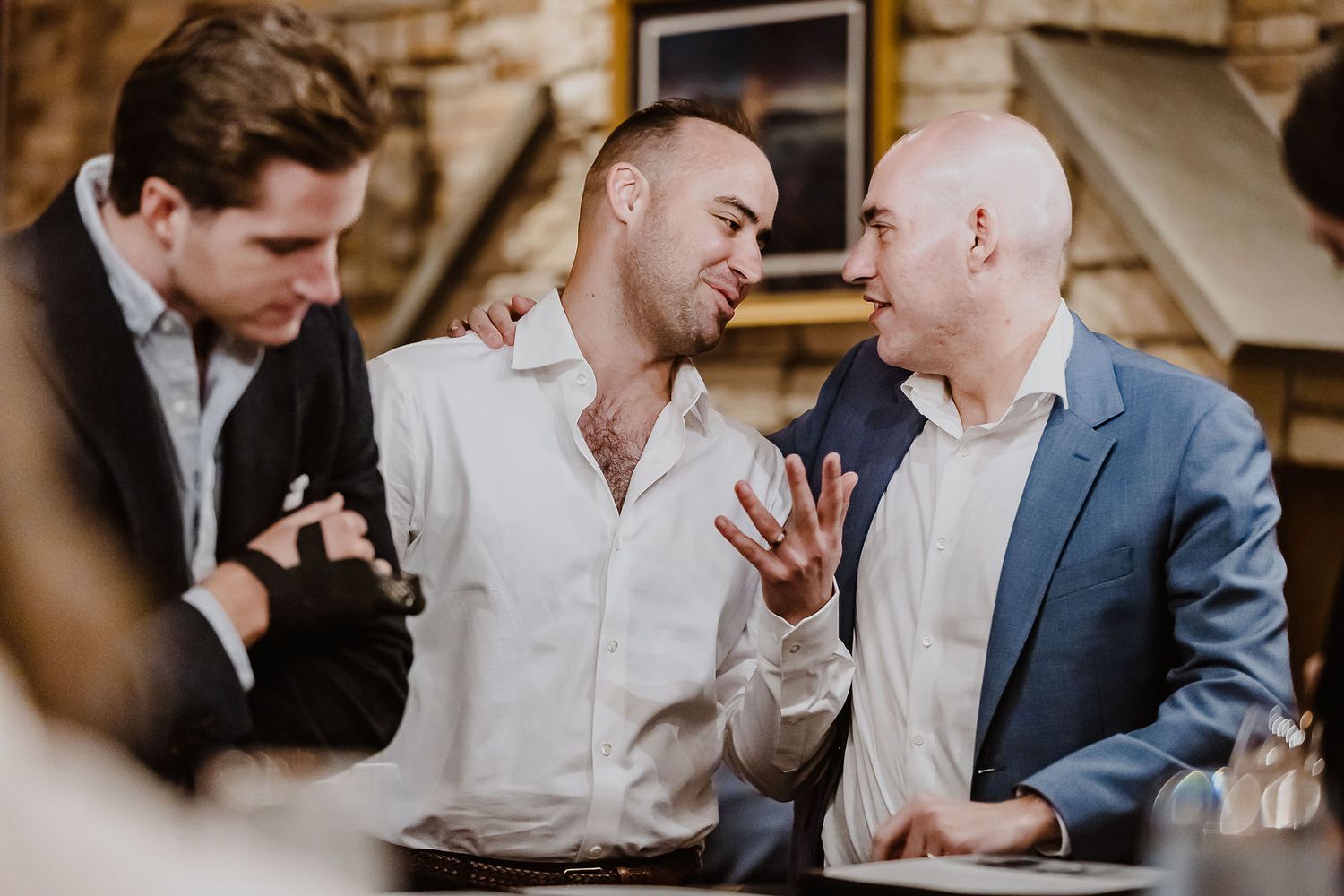 Three men in formal attire share a lively conversation and laughs at a wedding reception.