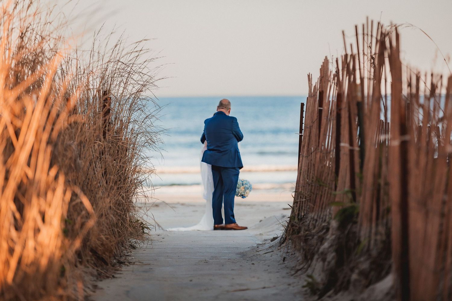 Solitary figure in blue suit walks toward ocean through a sandy beach path at sunset.