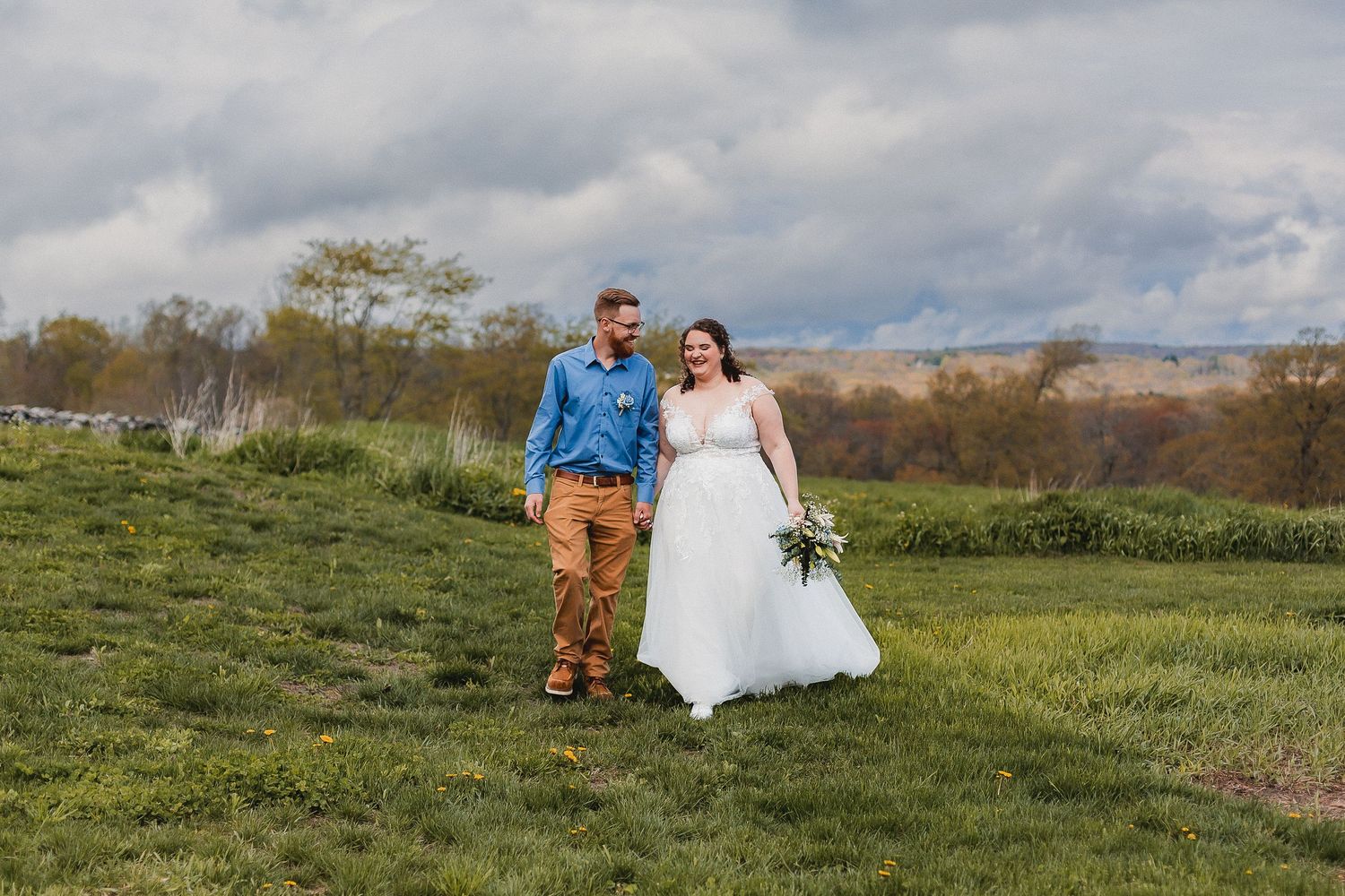 Bride in white gown and groom in blue shirt and tan pants walk hand in hand across a green field.