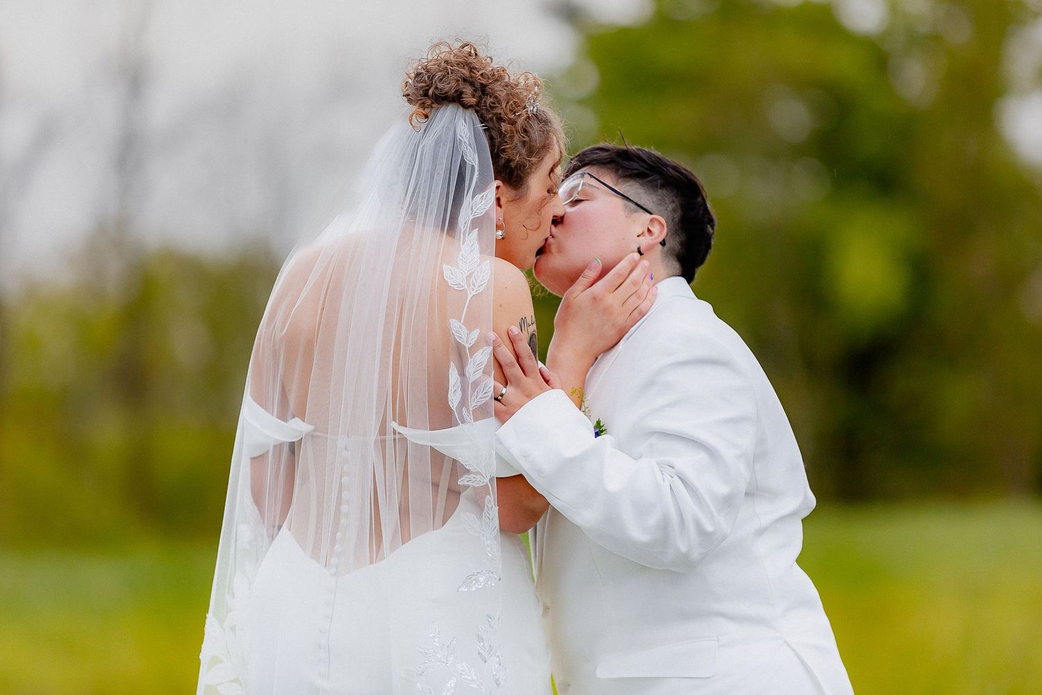 Two brides sharing a tender kiss outdoors, one in a white gown with veil on their wedding day.