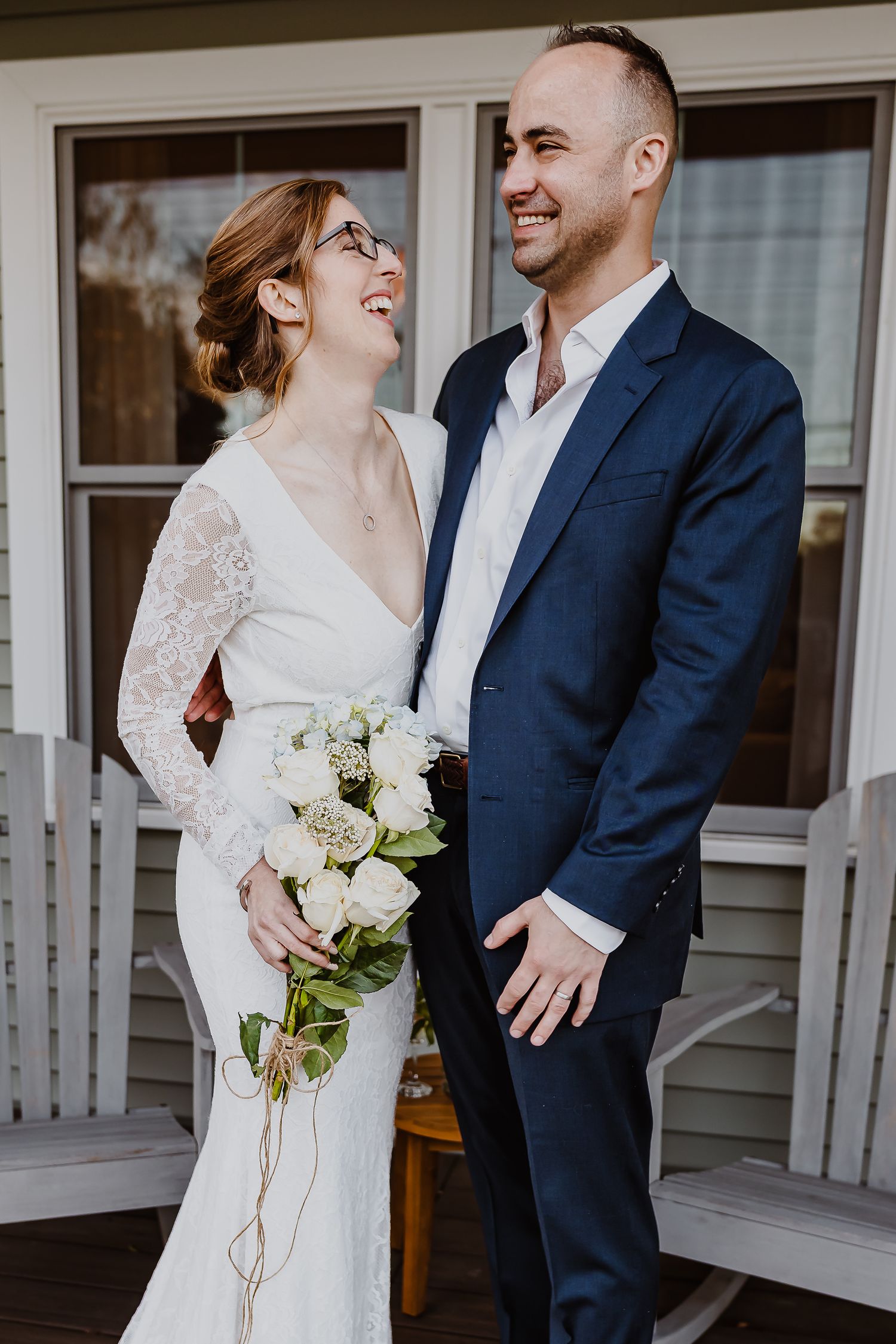 Smiling bride in lace gown holding white bouquet stands with groom in navy suit on a porch.