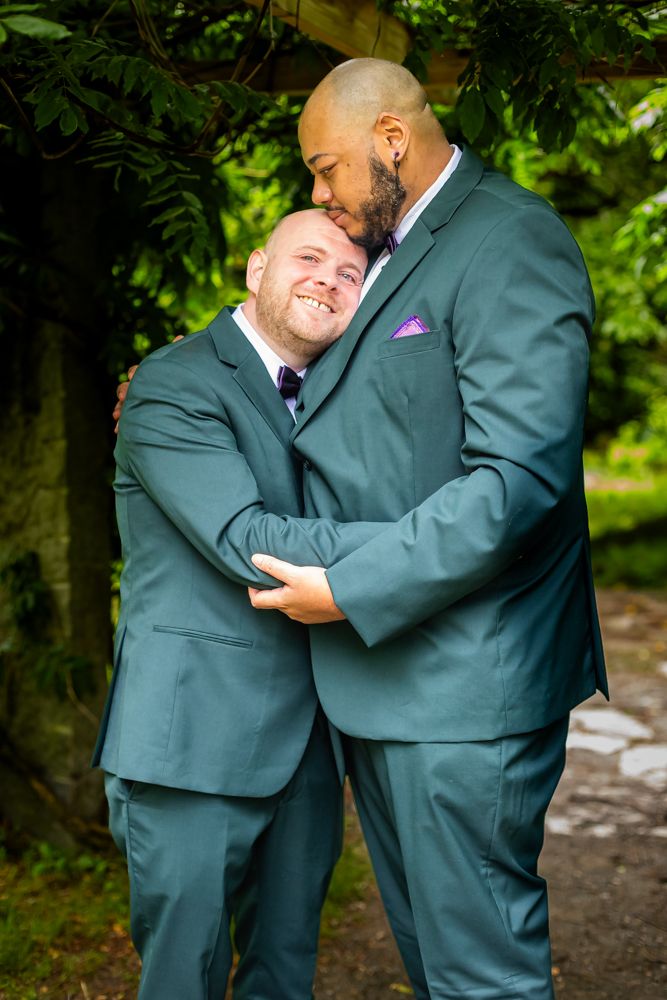 Two grooms in matching teal suits sharing an intimate embrace in a lush garden setting.