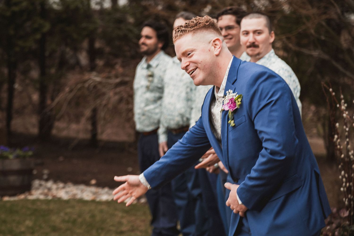 Smiling groom in a blue suit with floral boutonniere laughing with groomsmen outdoors.