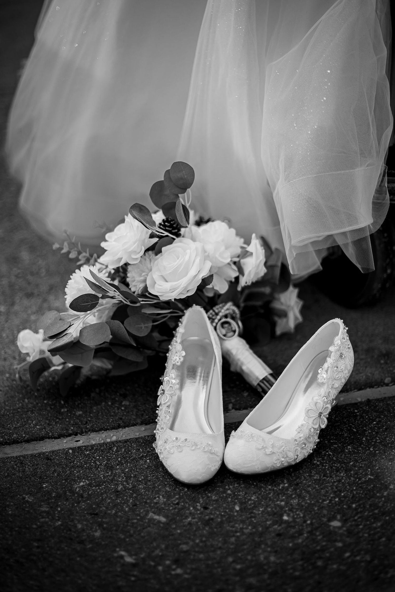 Black and white photo of white lace bridal heels beside a rose bouquet.