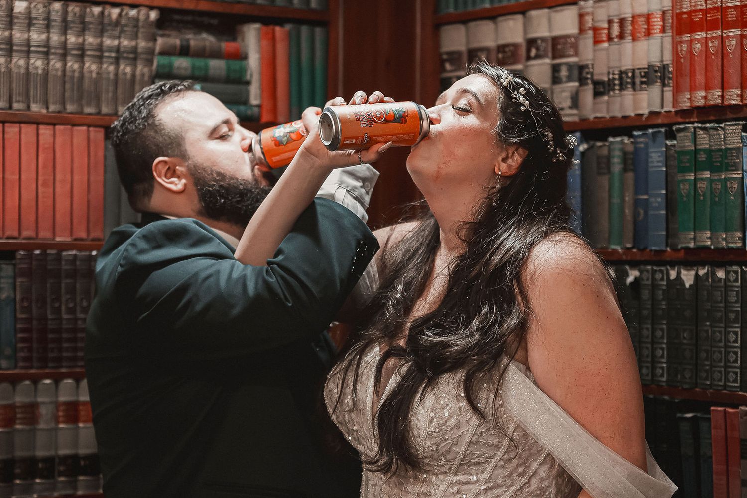 Bride and groom share a fun moment drinking from orange cans in front of bookshelves.