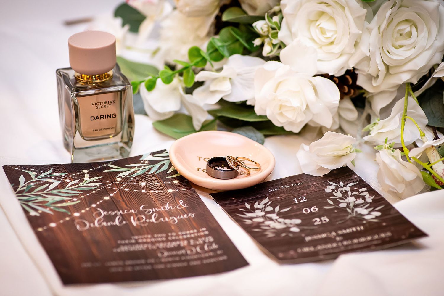 Wedding flat lay with wooden invitations, white roses, rings, and perfume bottle on white table.