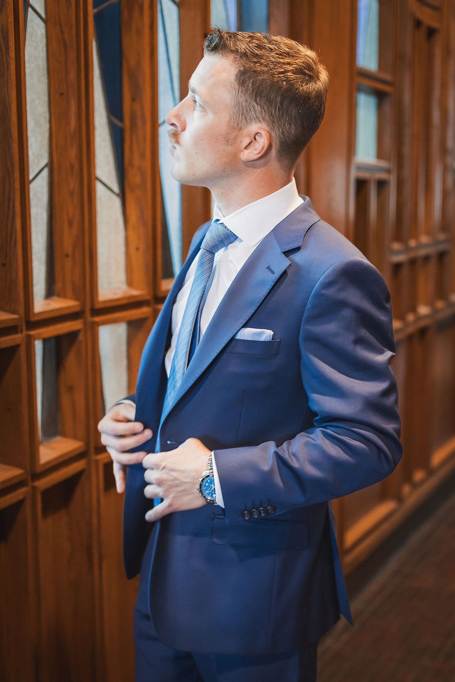 Dapper groom in navy blue suit adjusting jacket near ornate wooden paneled wall.