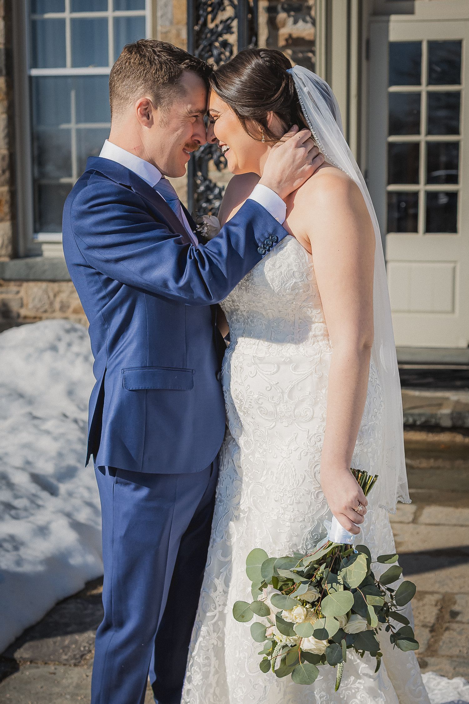Groom in blue suit tenderly touches brides face outside stone building in winter.