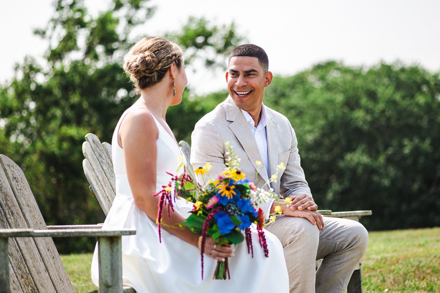 Bride and groom sitting on wooden chairs outdoors, sharing a joyful moment with colorful bouquet.