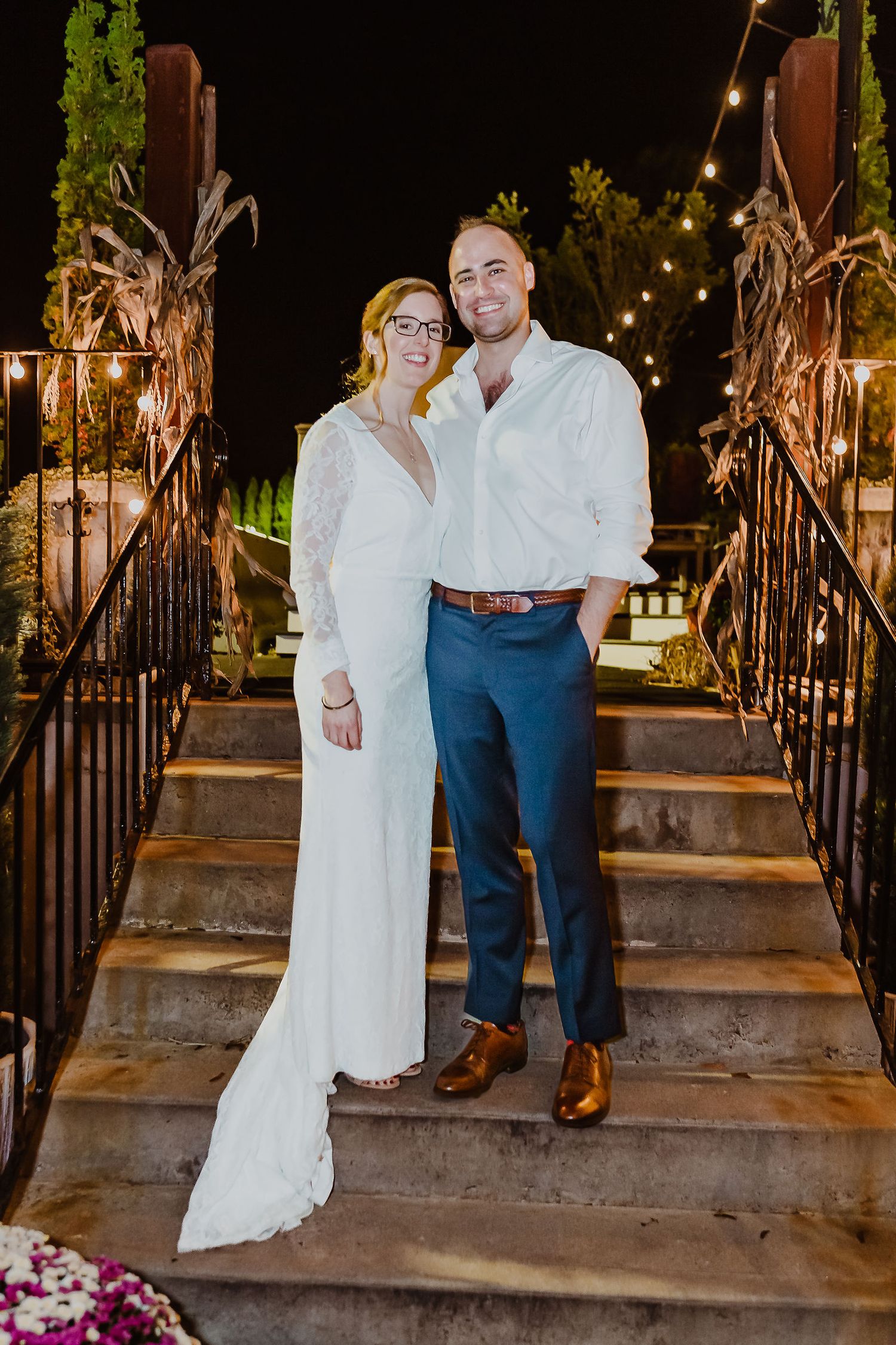 Newlywed couple smiling together on outdoor staircase with string lights at night.
