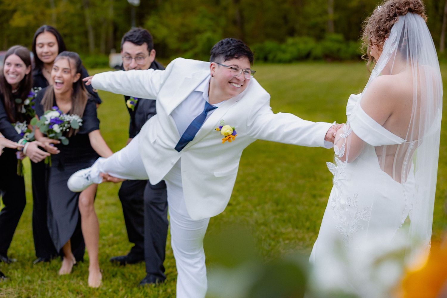 Joyful groom in white suit playfully kicking while holding bride's hand with wedding party behind.