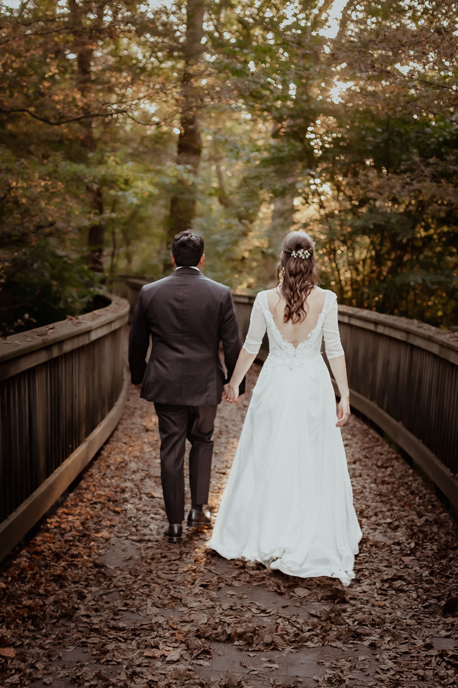 Bride and groom walk hand-in-hand across a leaf-covered wooden bridge surrounded by autumn trees.