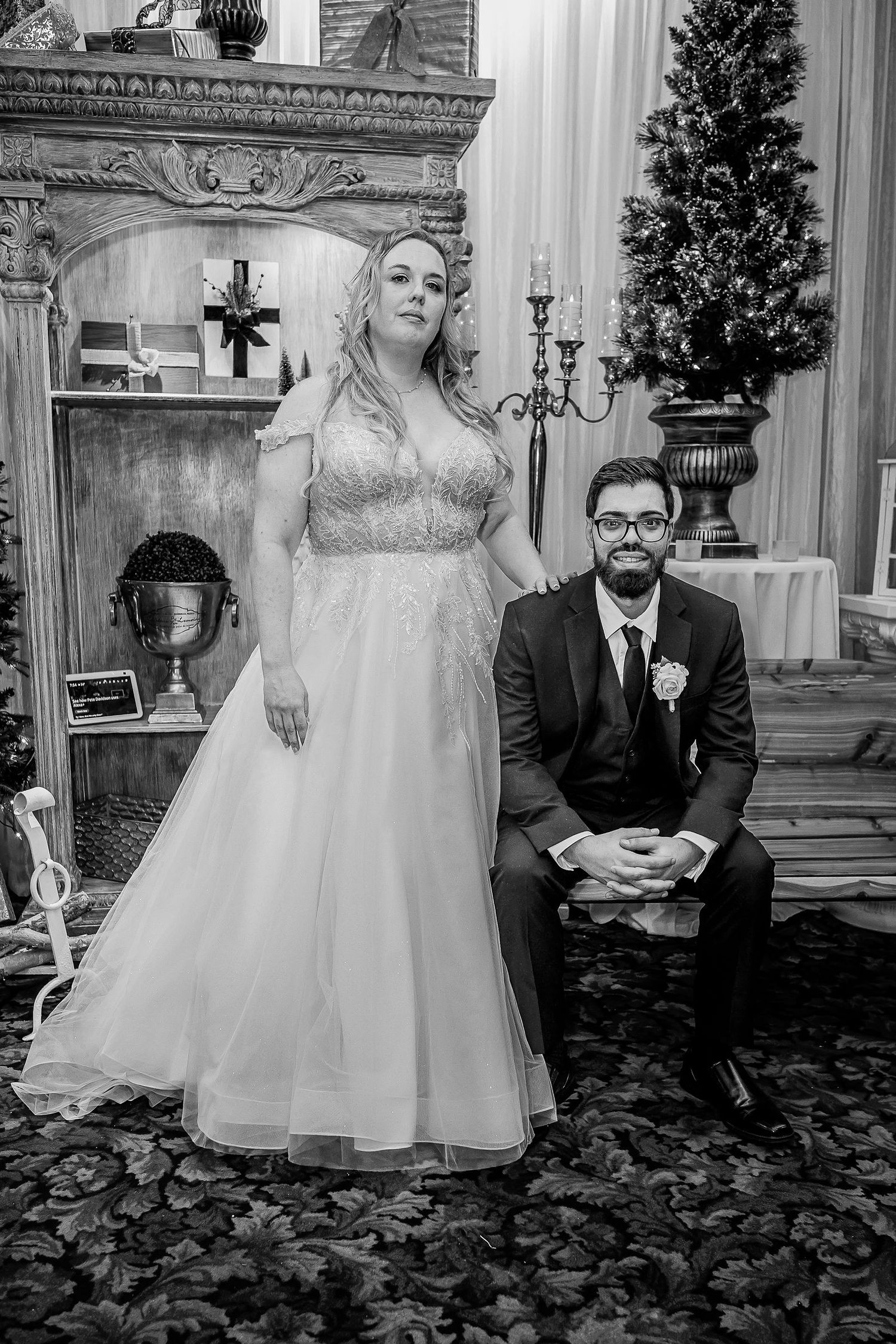 Black and white portrait of bride in ball gown and groom in suit posing by ornate fireplace.