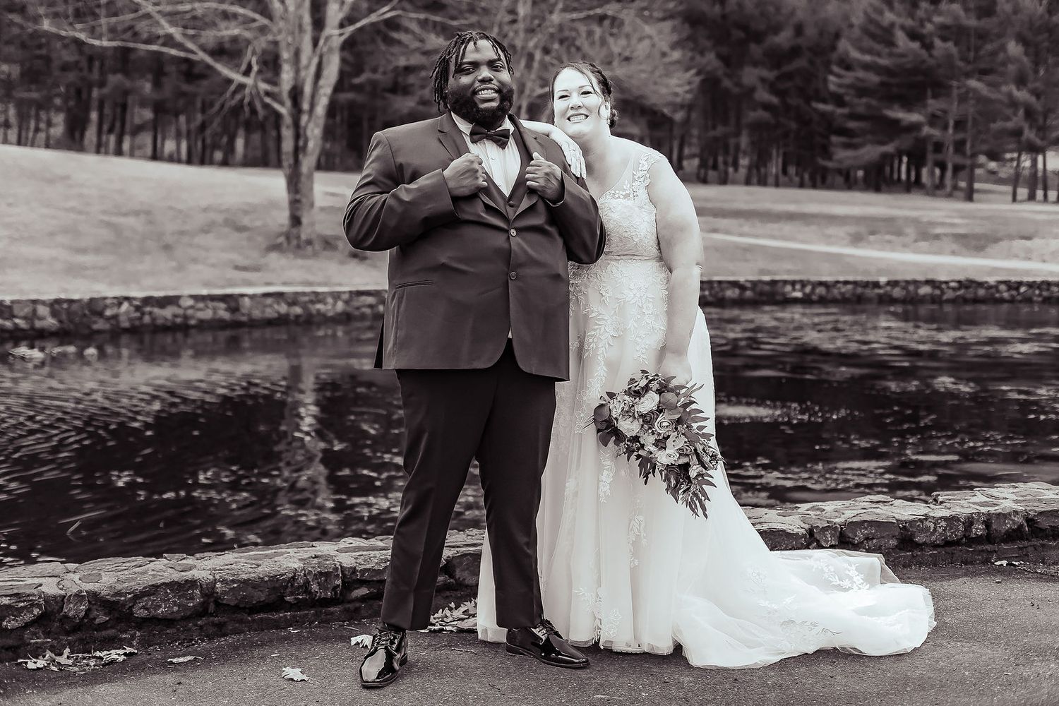 Black and white photo of newlywed couple posing playfully near scenic pond.