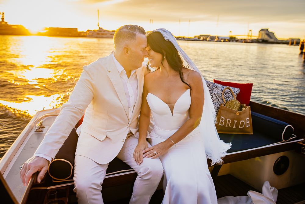 Bride and groom sit closely on a boat at golden sunset, with a Bride pillow nearby.