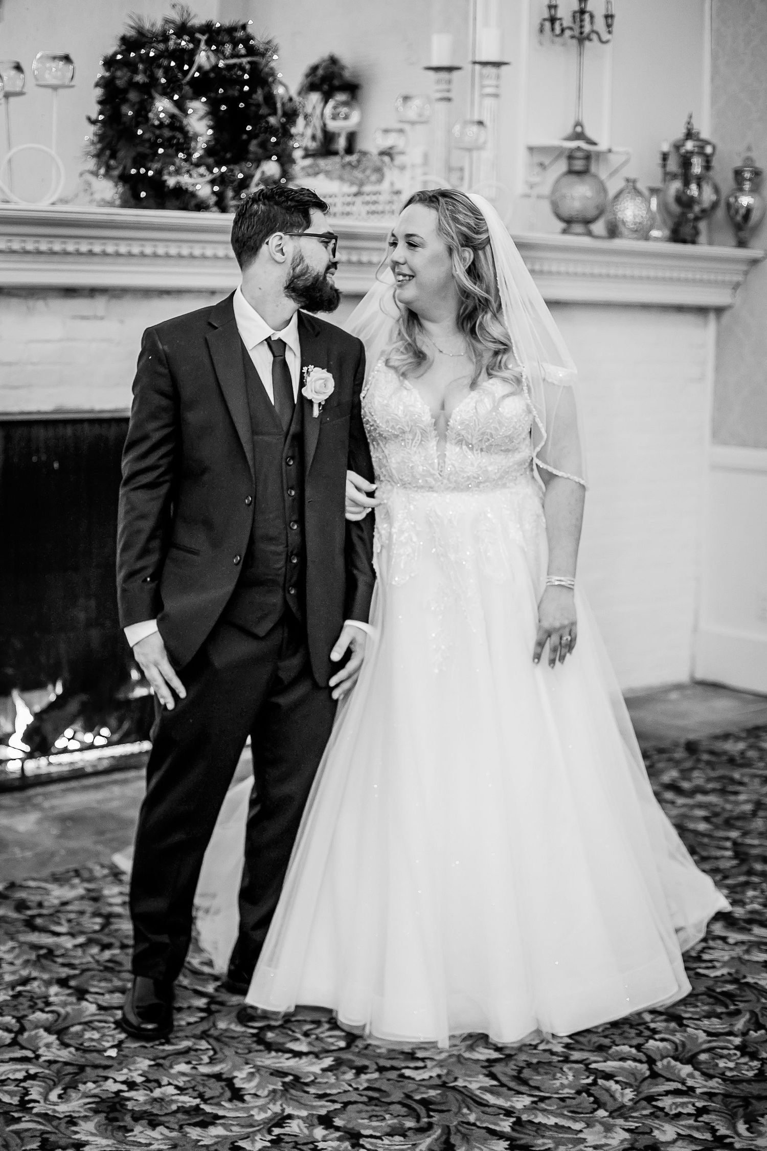 Black and white photo of smiling bride and groom standing together near fireplace at wedding.